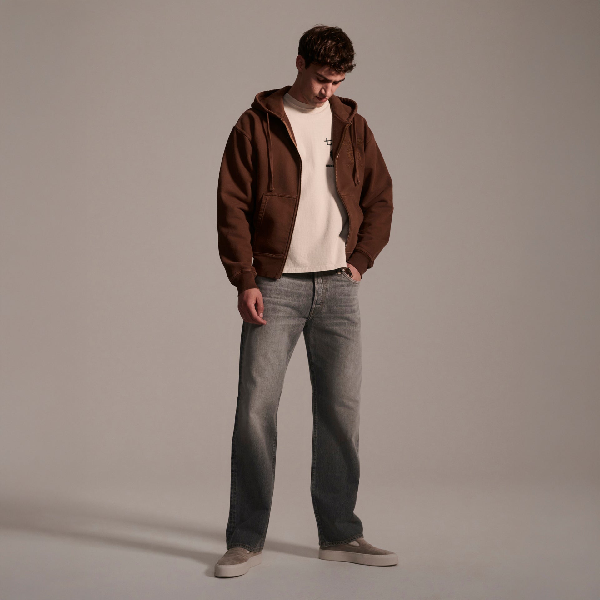 A person stands in a studio wearing a brown zip-up hoodie over a white t-shirt, T7C’s The Jack Jean in Cloud, and beige shoes, looking down with hands in jacket pockets against a neutral background.