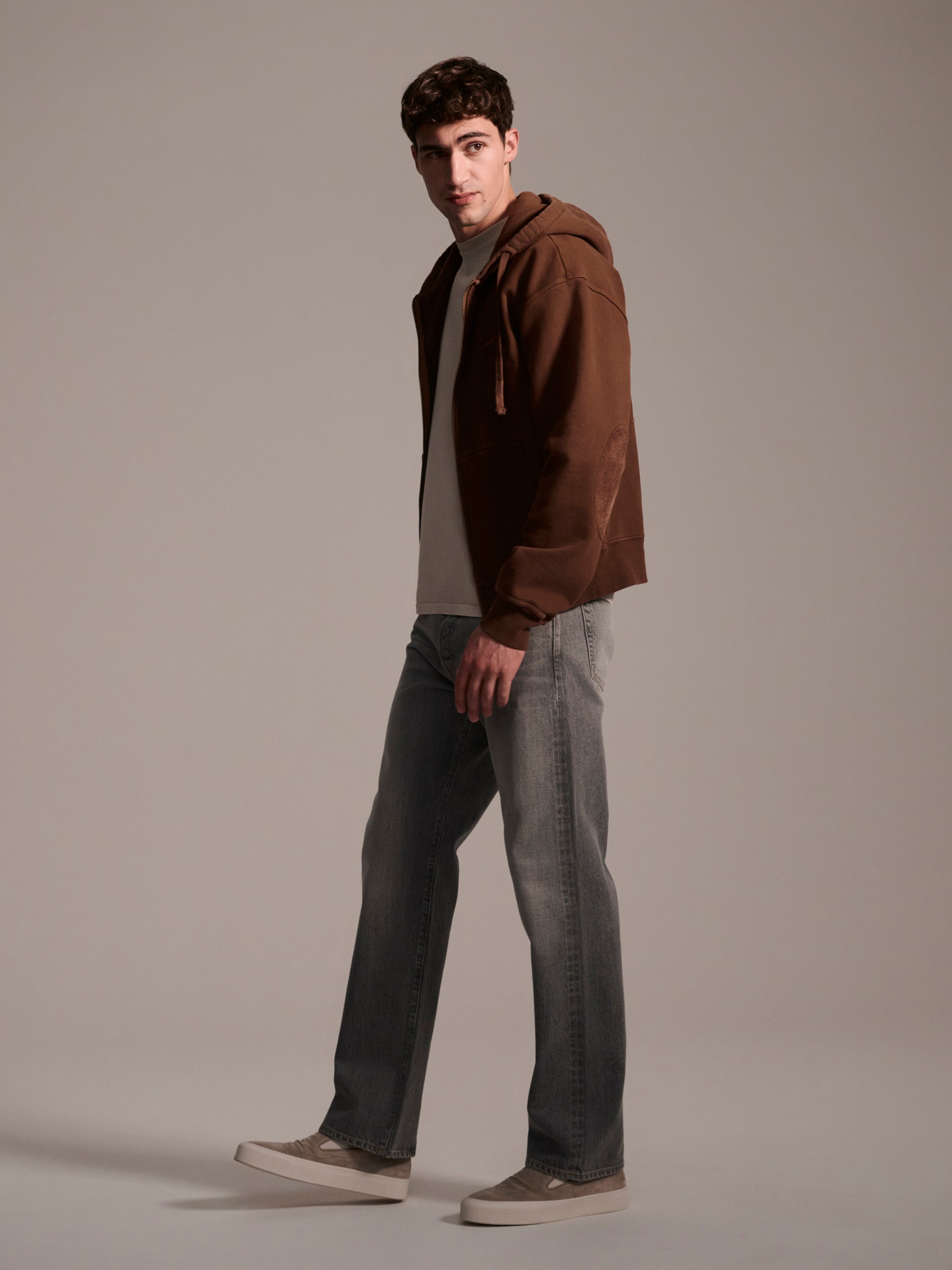 A man stands against a plain background wearing a brown hoodie, beige t-shirt, T7Cs The Jack Jean in Cloud, and beige sneakers, looking slightly to his right with one foot forward.