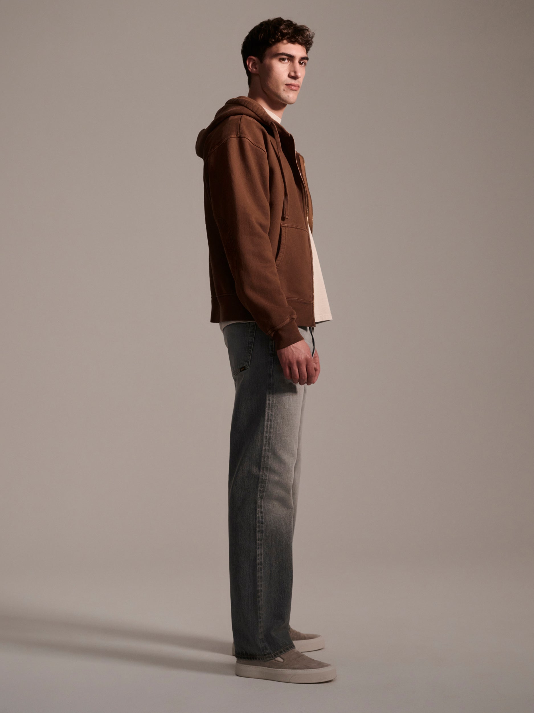 A young man stands sideways against a plain background, wearing a brown hoodie, beige shoes, and T7C’s The Jack Jean in Cloud. He looks ahead with a neutral expression.