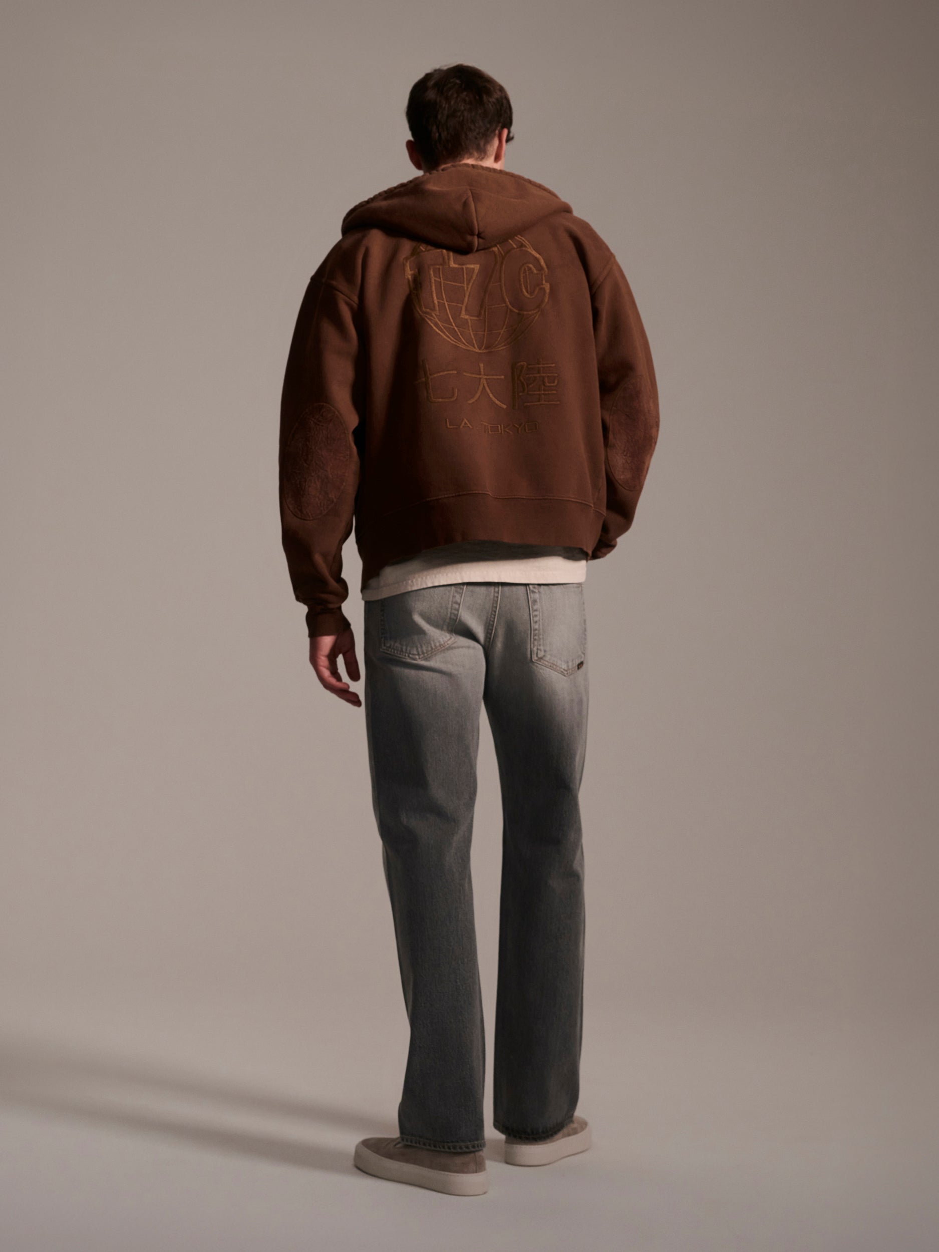 A person stands facing away, wearing T7C’s The Jack Jean in Cloud—stone-washed light gray denim—paired with a brown hooded sweatshirt with gold lettering and beige shoes against a plain background.