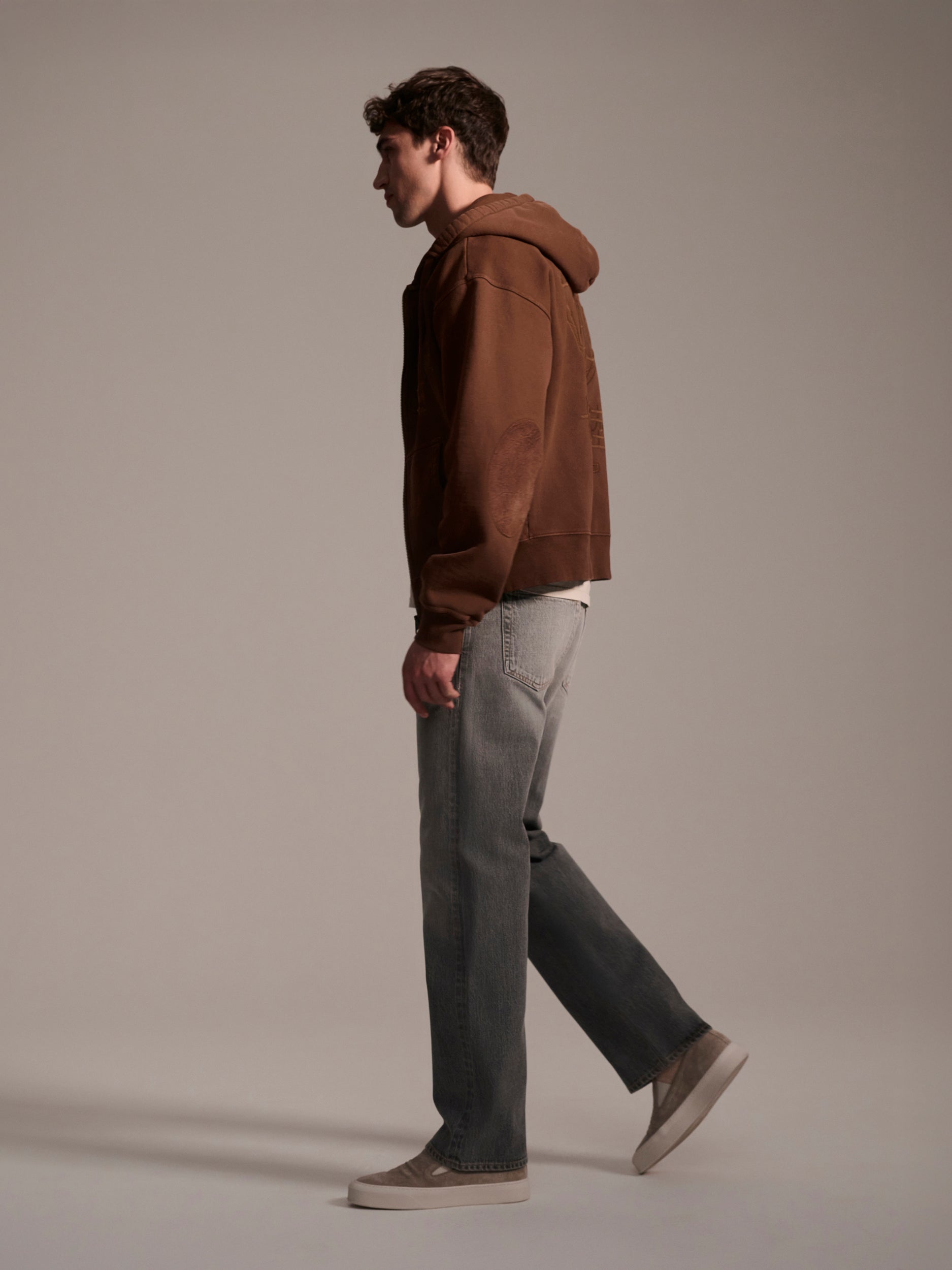 A man stands in profile against a plain background, wearing a brown hoodie, T7Cs The Jack Jean in Cloud, and beige sneakers.