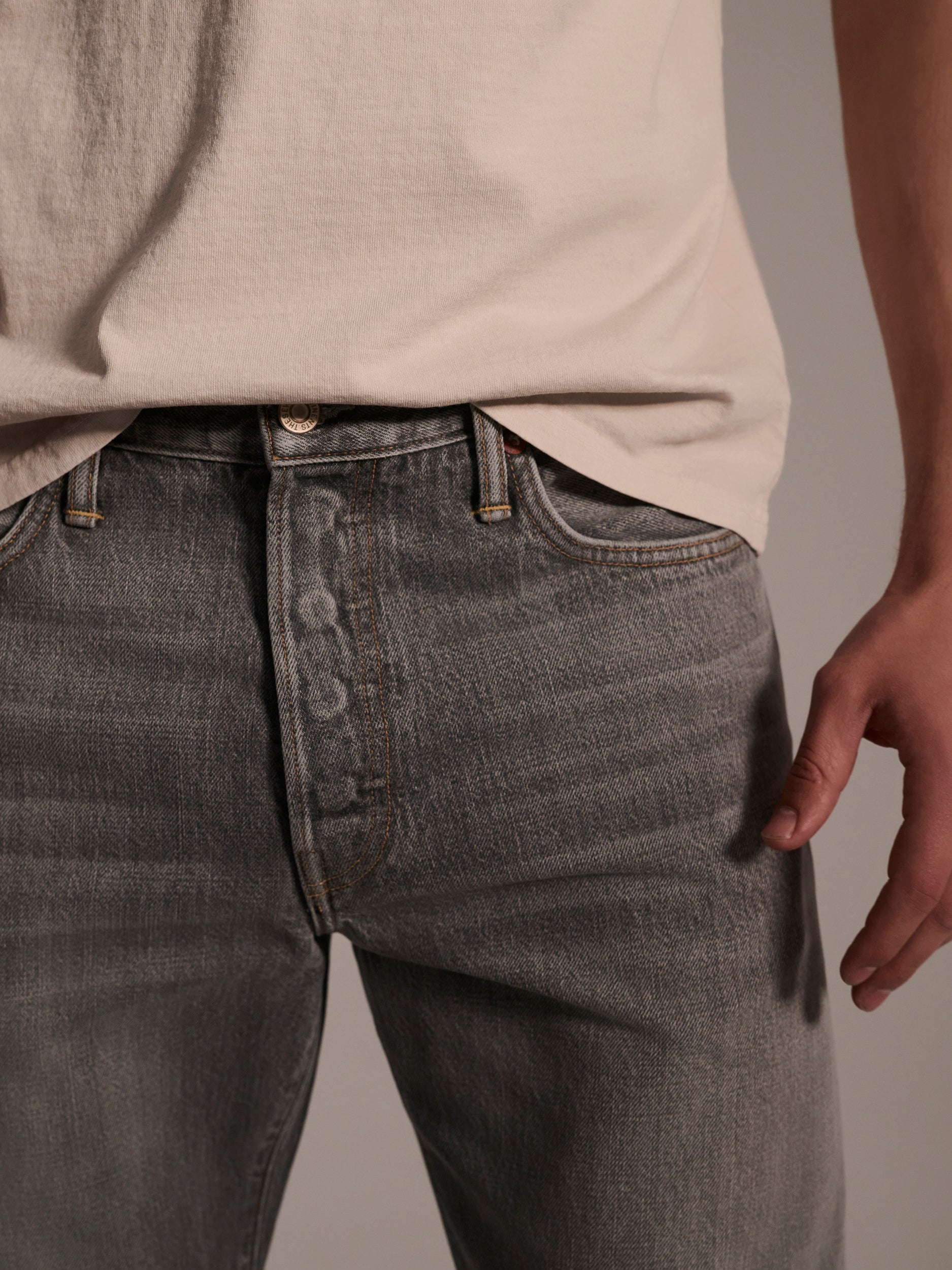 A person wearing a beige t-shirt and The Jack Jean in Cloud by T7C stands with one hand by their side, shown from the waist down against a plain background.
