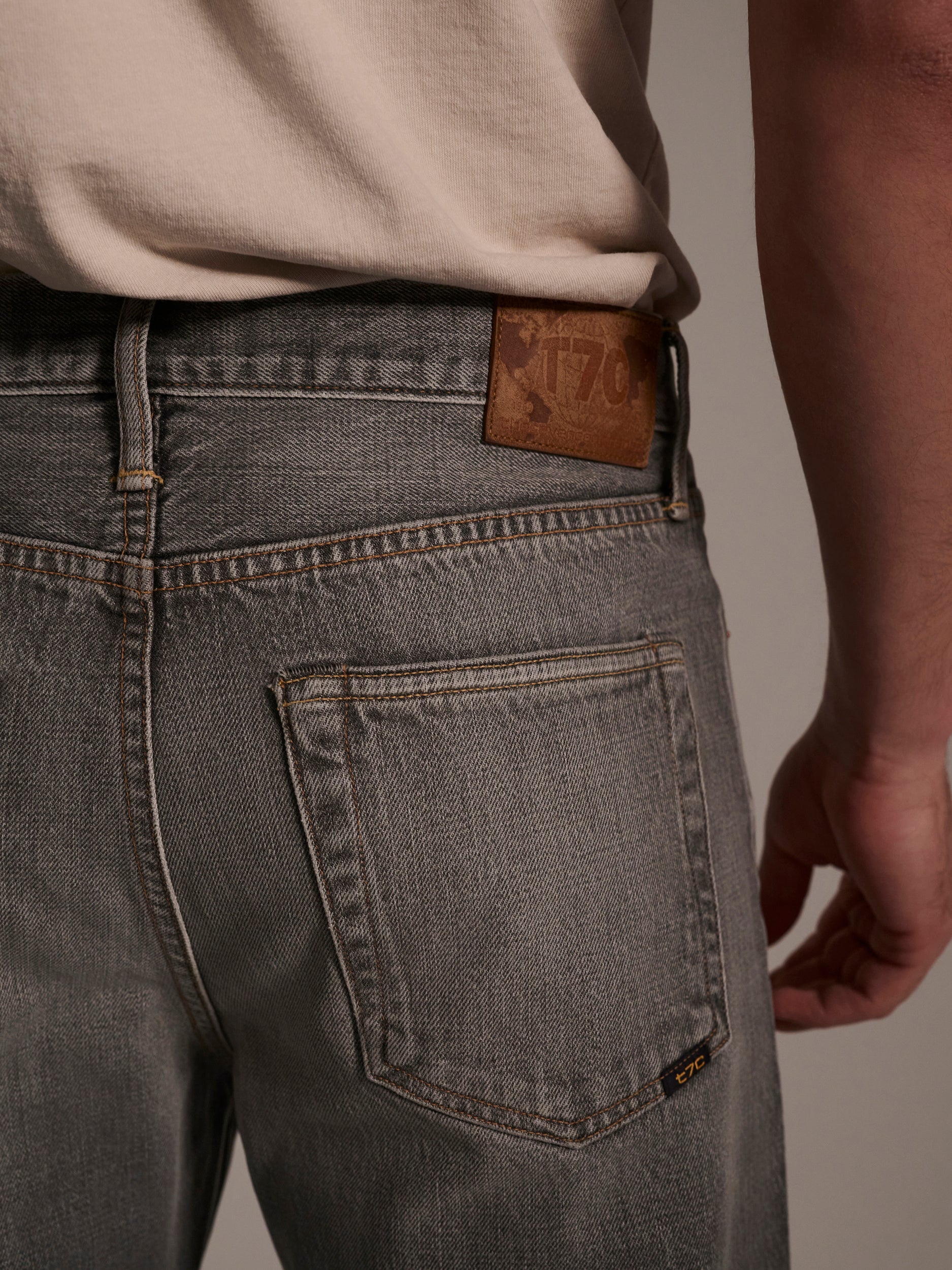 Close-up of a person wearing T7Cs The Jack Jean in Cloud—stone-washed light gray jeans crafted from organic Turkish cotton, featuring a leather patch on the waistband and a back pocket. A light shirt is tucked in.