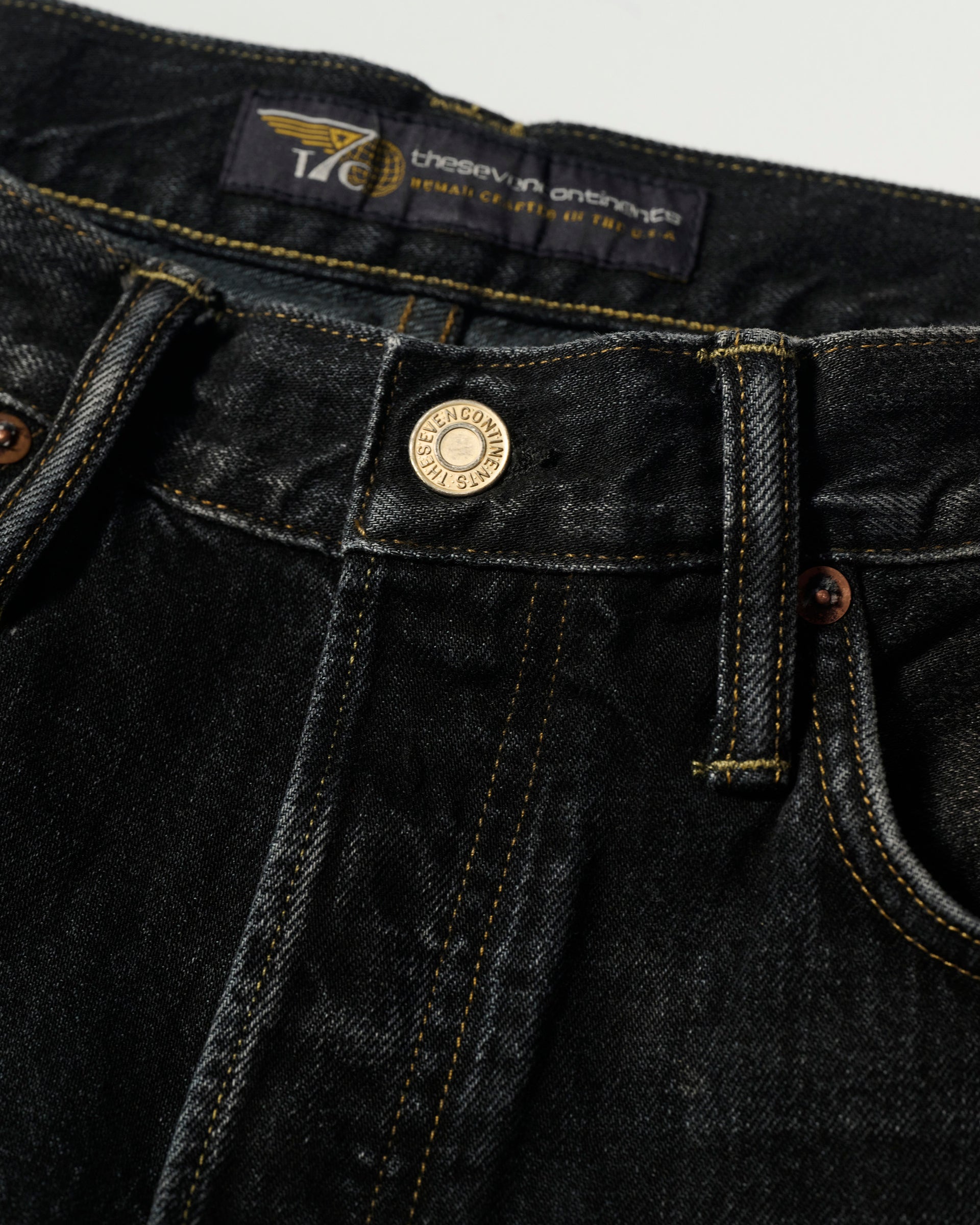 A close-up of the waistband and front button of The Jack Jean In Kobe Black by T7C, featuring yellow stitching, belt loops, a branded metal button, and a visible label inside the waistband.