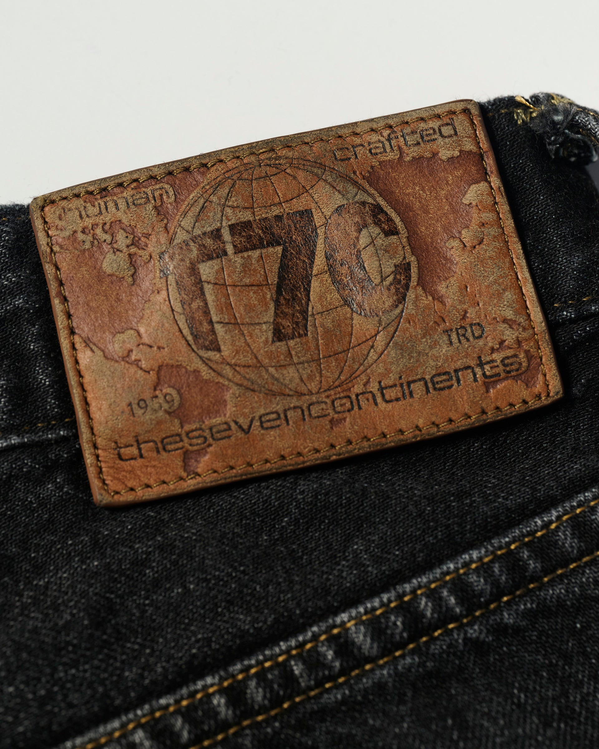 A close-up of the brown leather patch on T7Cs The Jack Jean In Kobe Black, featuring a globe design, T7C, the seven continents, human crafted, and TRD 1999 on black Japanese selvedge denim.