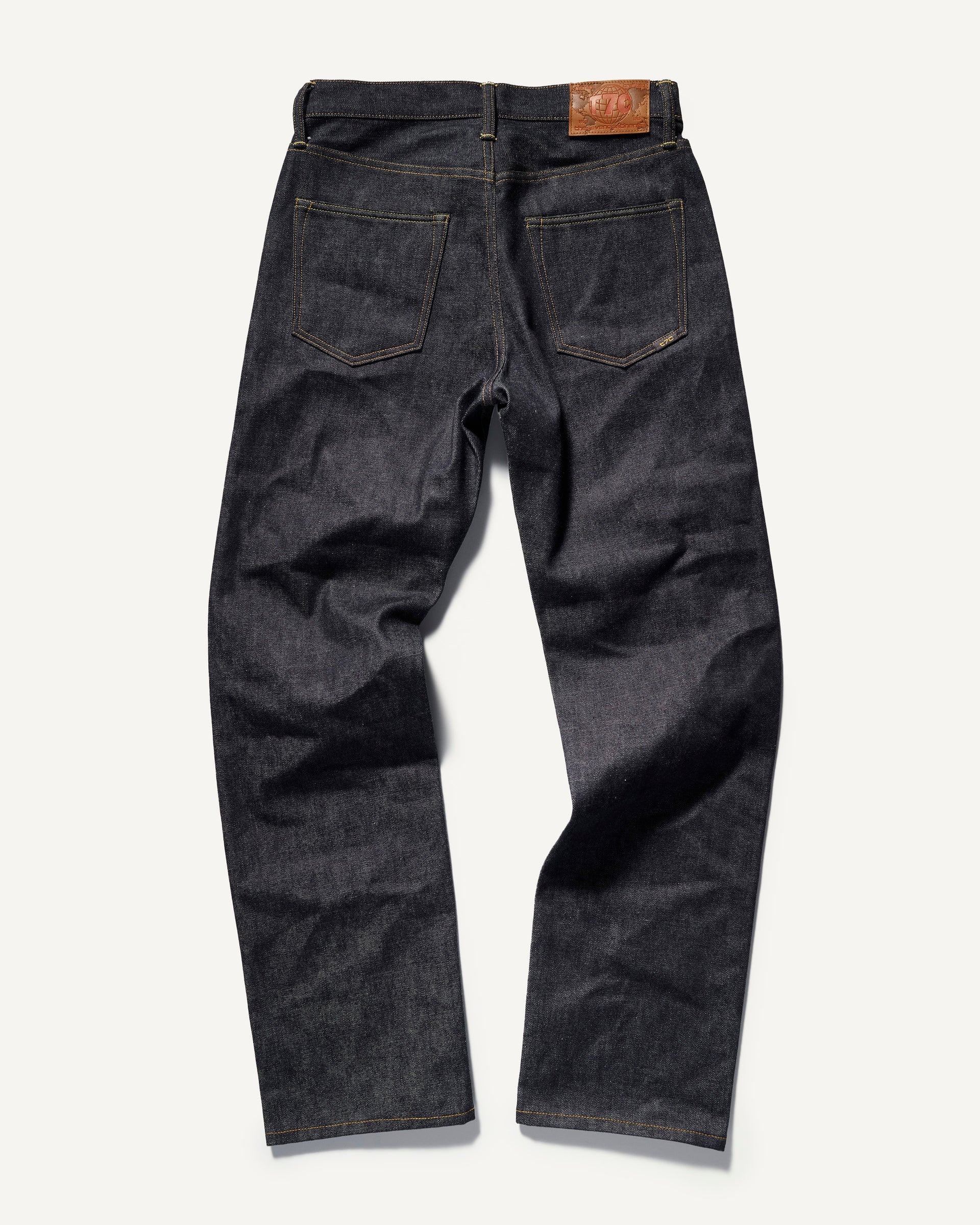 The Jack Jean In Yokohama Raw by T7C The Seven Continents is shown from the back on a white background, featuring two pockets, yellow stitching, and a brown leather patch. Made from rope-dyed indigo selvedge denim.