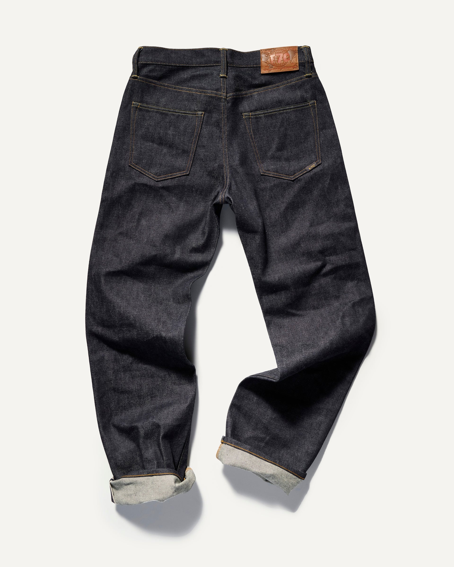 The Jack Jean In Yokohama Raw by T7C The Seven Continents: dark blue, rope-dyed Japanese denim with rolled cuffs, two back pockets, yellow stitching, and a brown leather waistband patch. Seen from the back.