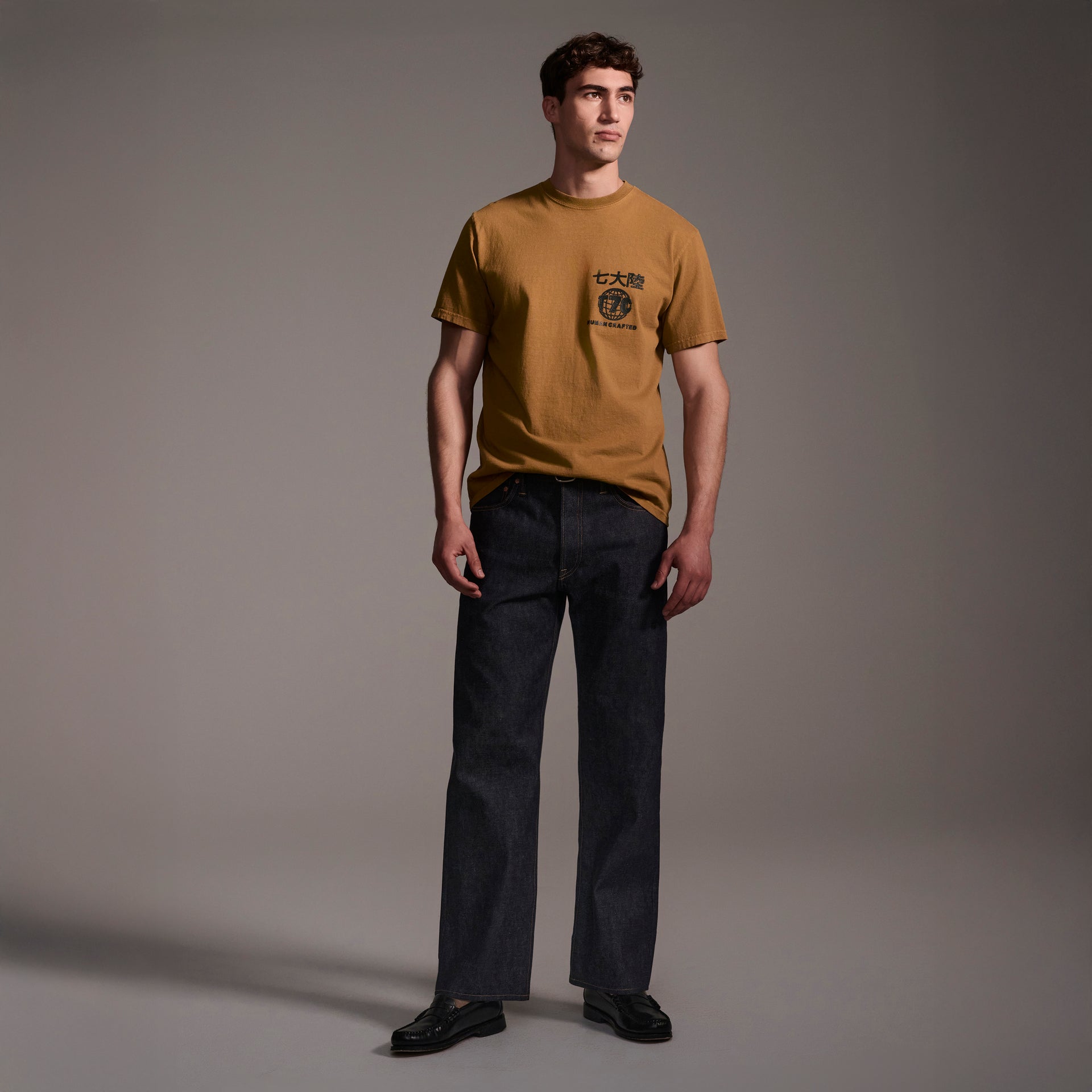 A man stands in a studio wearing a mustard yellow graphic tee, black shoes, and The Jack Jean In Yokohama Raw by T7C The Seven Continents. The background is plain gray with soft shadows.