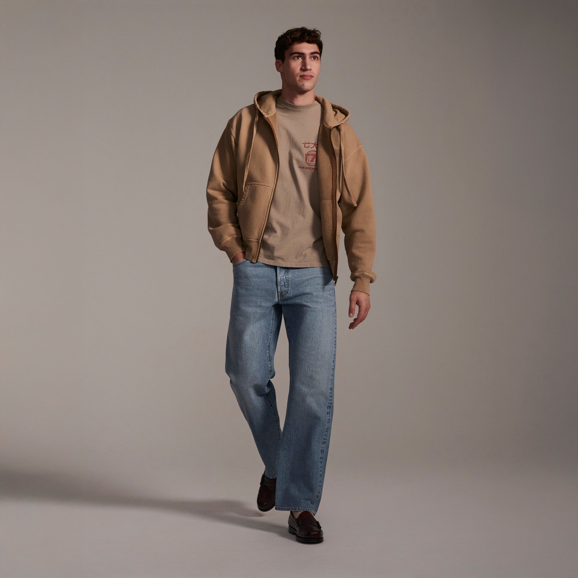 A man confidently walks against a neutral background wearing T7C The Seven Continents’ The Jack Jean in Zuma, tan hoodie, beige graphic tee, and dark brown shoes.