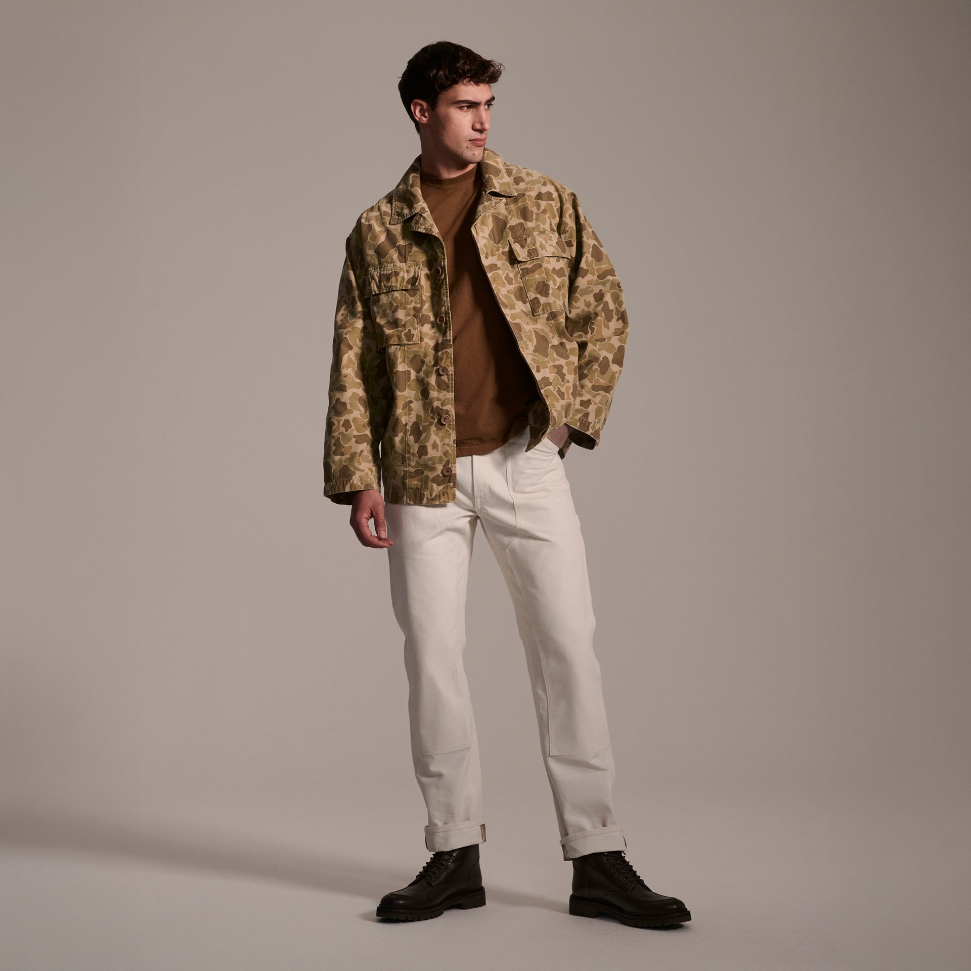 A man in a studio wears a camouflage jacket, brown turtleneck, and The Sid Pant in Chalk Raw by T7C The Seven Continents with black boots—modern workwear style, gazing to the side with one hand in his pocket.