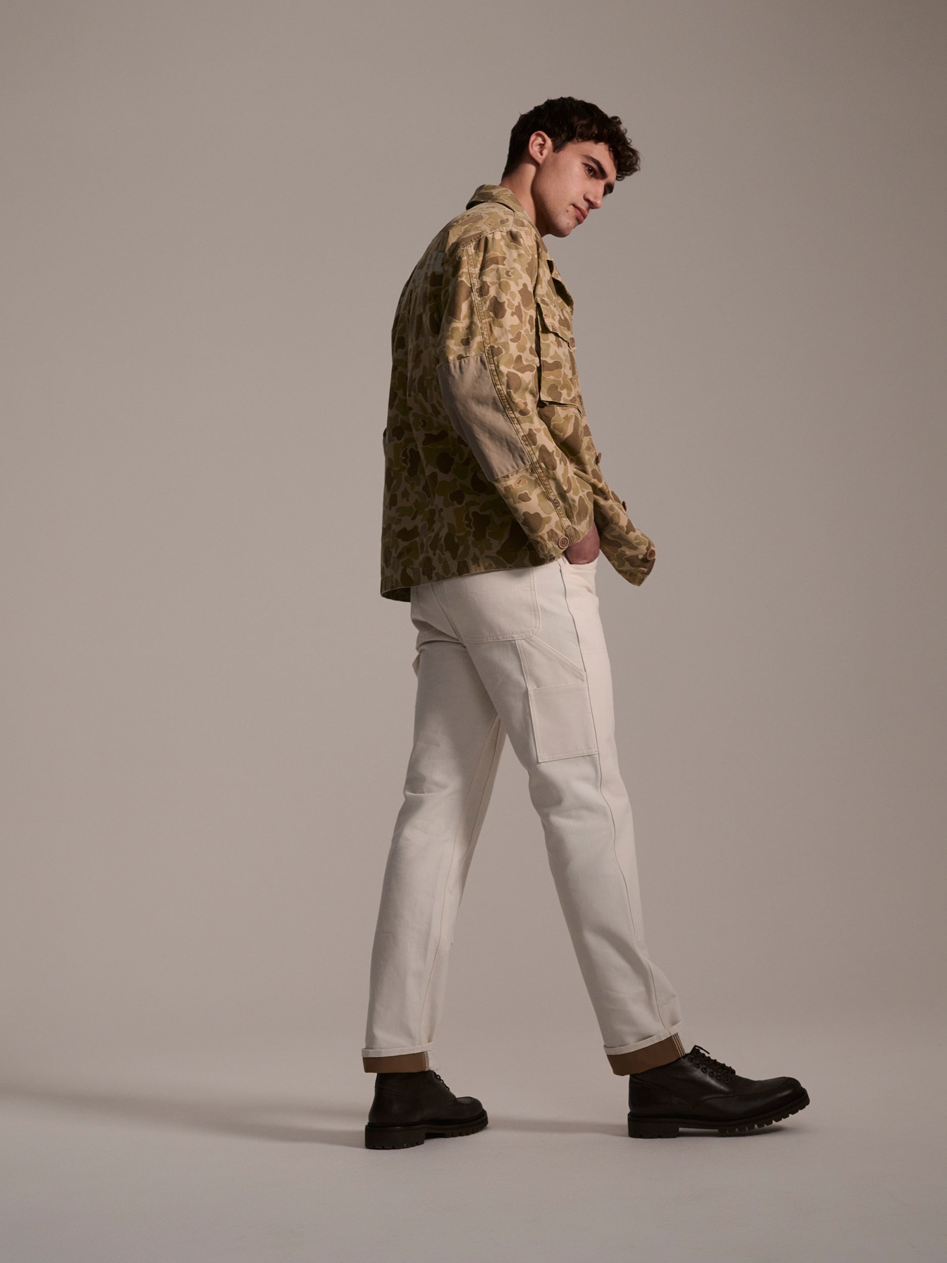 A man with dark hair stands in a studio, hands in pockets, wearing T7C The Seven Continents Sid Pant in Chalk Raw and a beige camo jacket. The plain background highlights his modern workwear style.