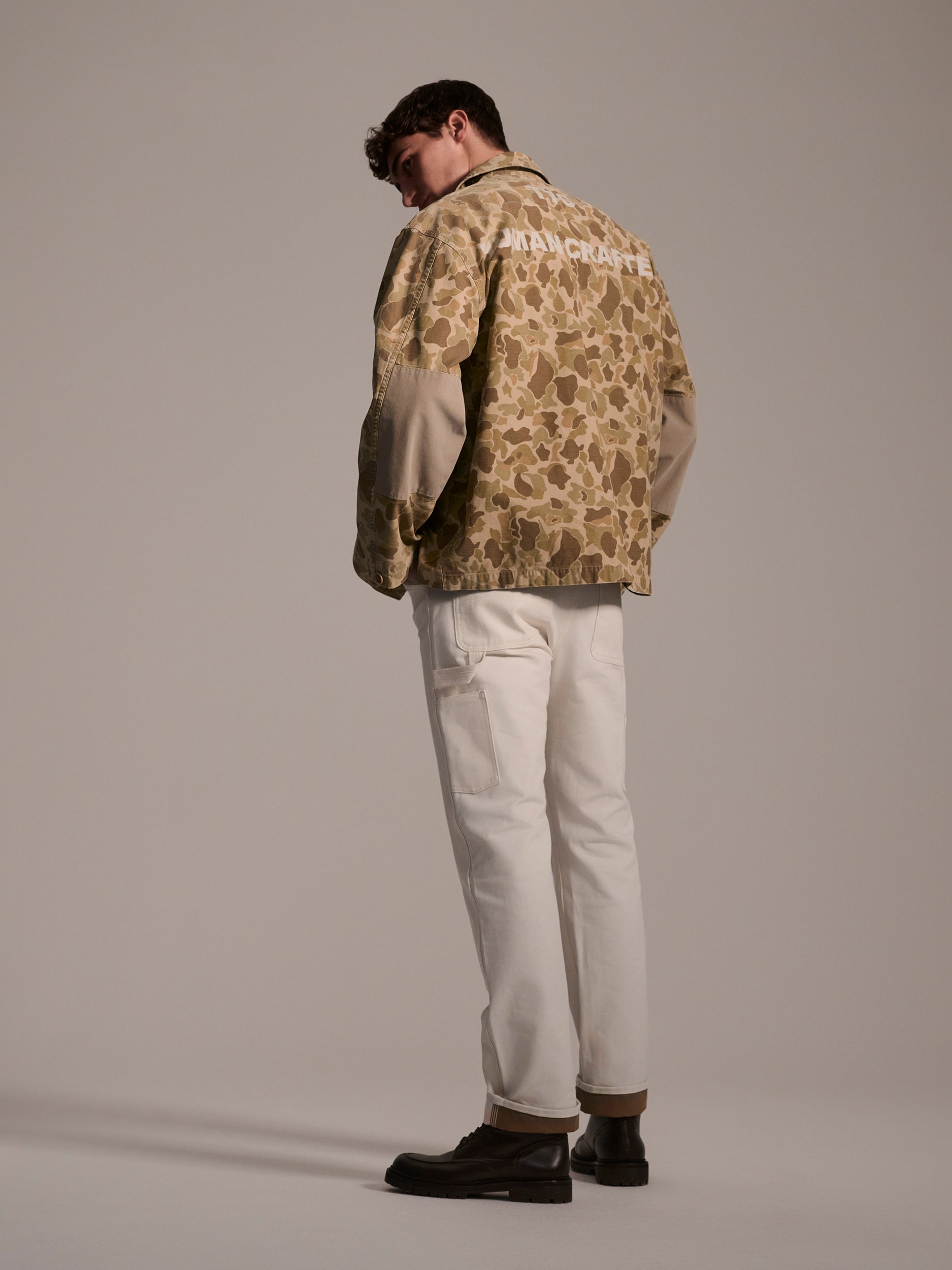 A man stands facing away, wearing a tan camouflage workwear jacket, T7C The Seven Continents Sid Pant in Chalk Raw, and dark shoes against a plain background.