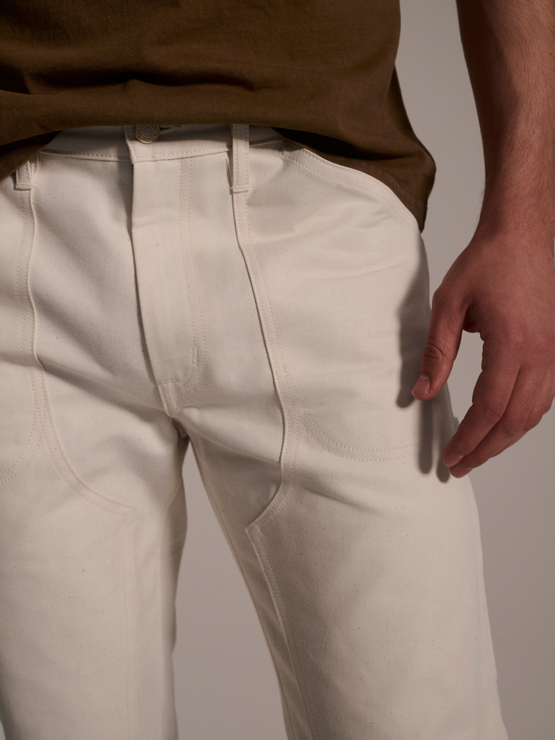 A person wearing T7C The Seven Continents The Sid Pant in Chalk Raw and a brown shirt, shown waist to mid-thigh with their right arm by their side—a modern update of classic carpenter workwear.