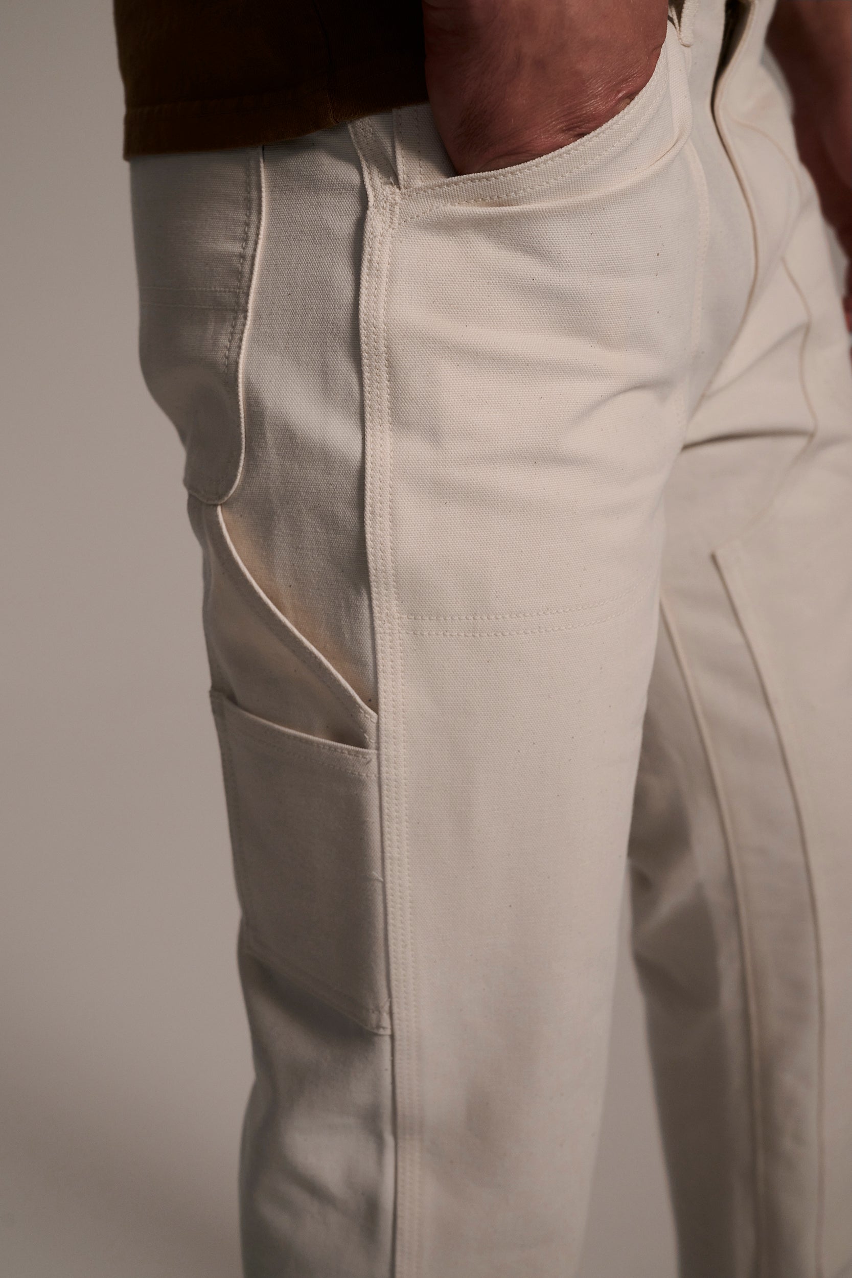 Close-up of a person wearing T7C The Seven Continents The Sid Pant in Chalk Raw, a relaxed-fit, cream-colored carpenter style with visible stitching, side pocket, classic detailing, and their right hand in the front pocket.