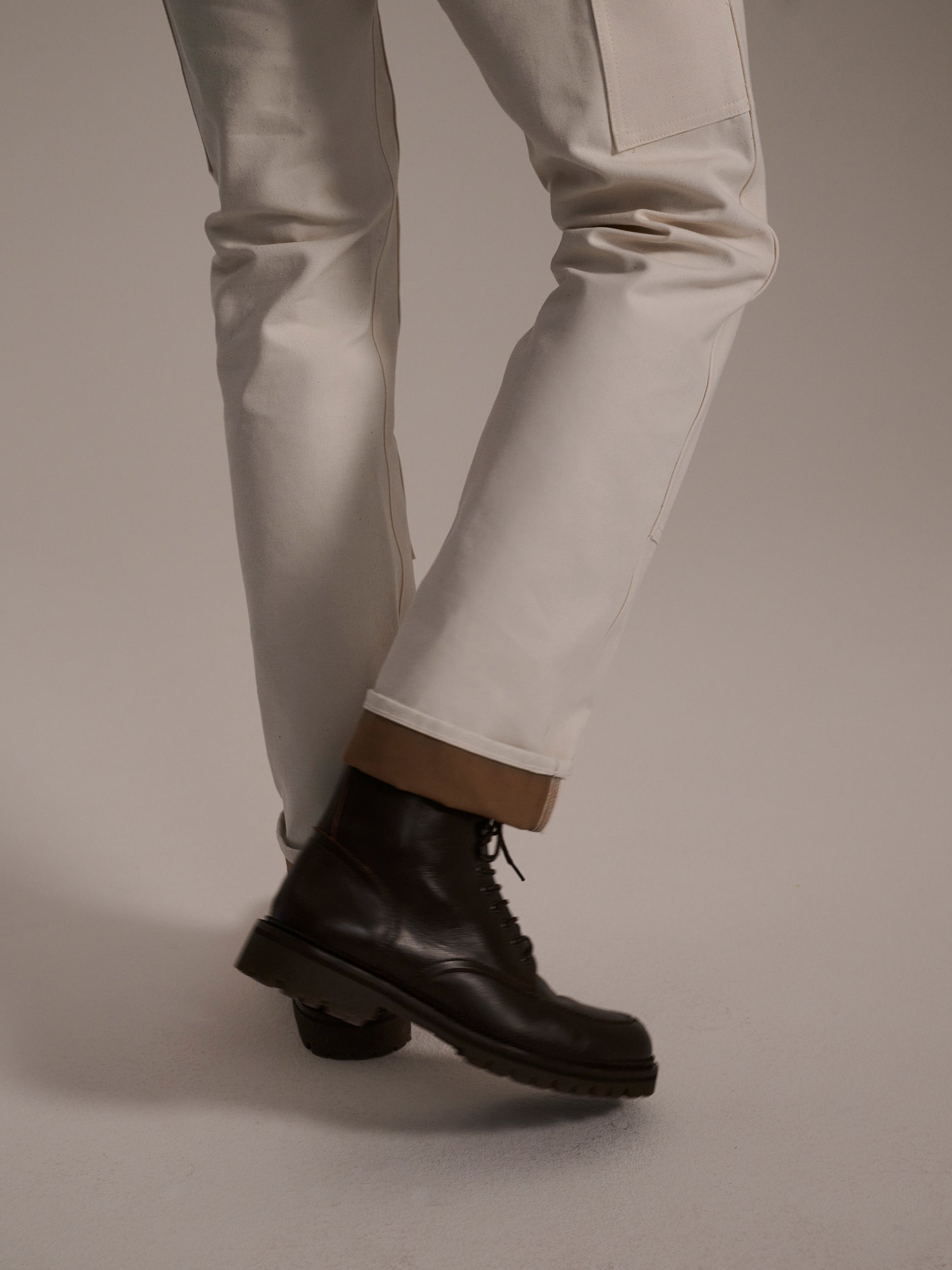 Wearing The Sid Pant in Chalk Raw by T7C The Seven Continents and black lace-up boots, a person stands with one leg crossed in front of the other against a plain background.