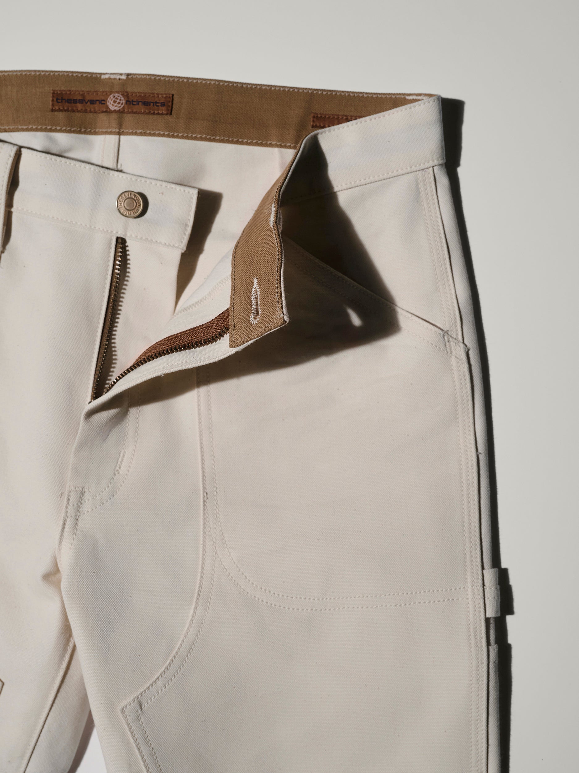 Close-up of The Sid Pant in Chalk Raw by T7C The Seven Continents, featuring an off-white hue, brown waistband, visible zipper and button closure, and detailed stitching against a light background.