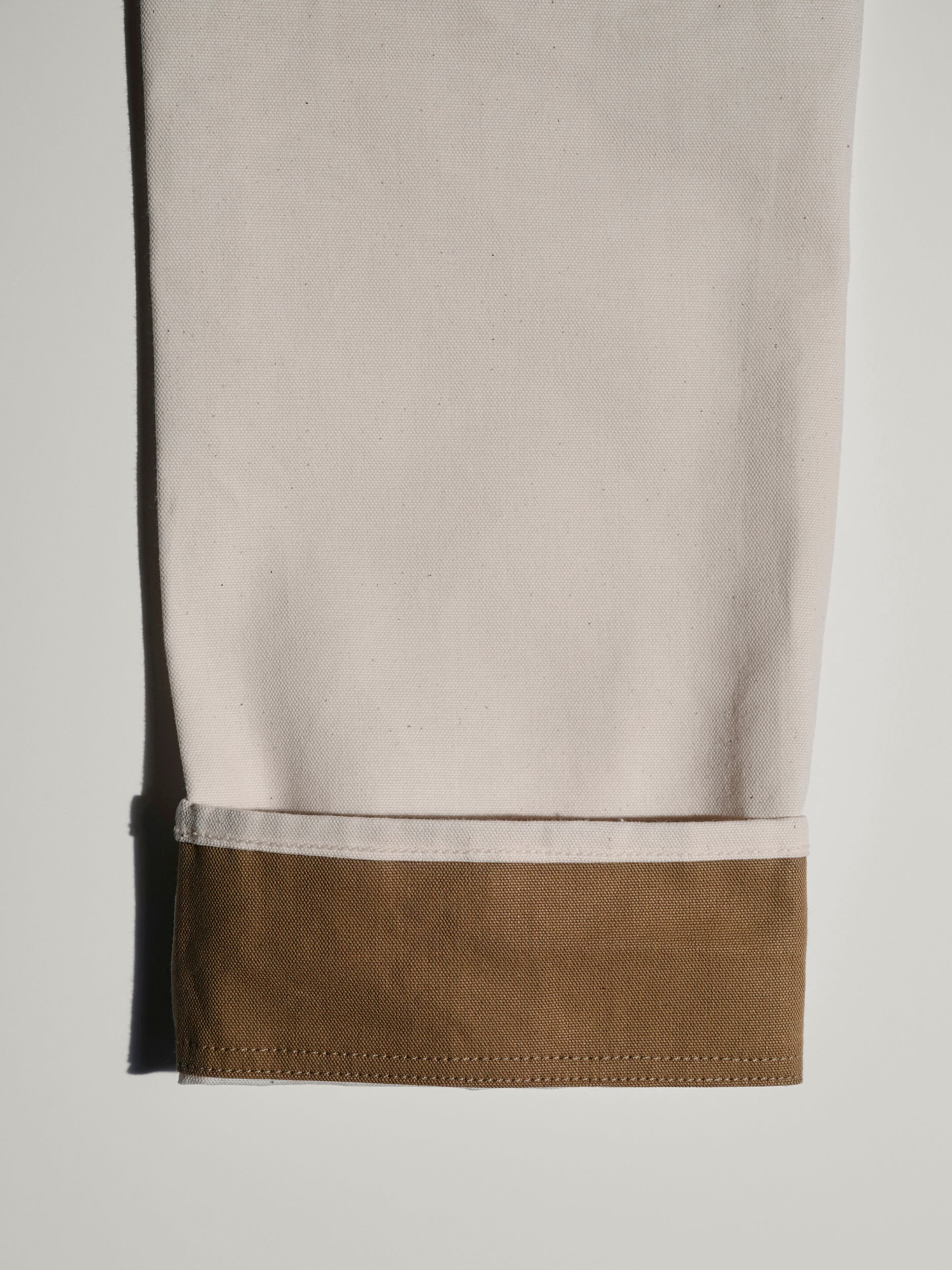 A close-up of the lower part of The Sid Pant in Chalk Raw by T7C The Seven Continents, featuring a wide brown cuff at the hem—versatile workwear style laid flat on a light surface.