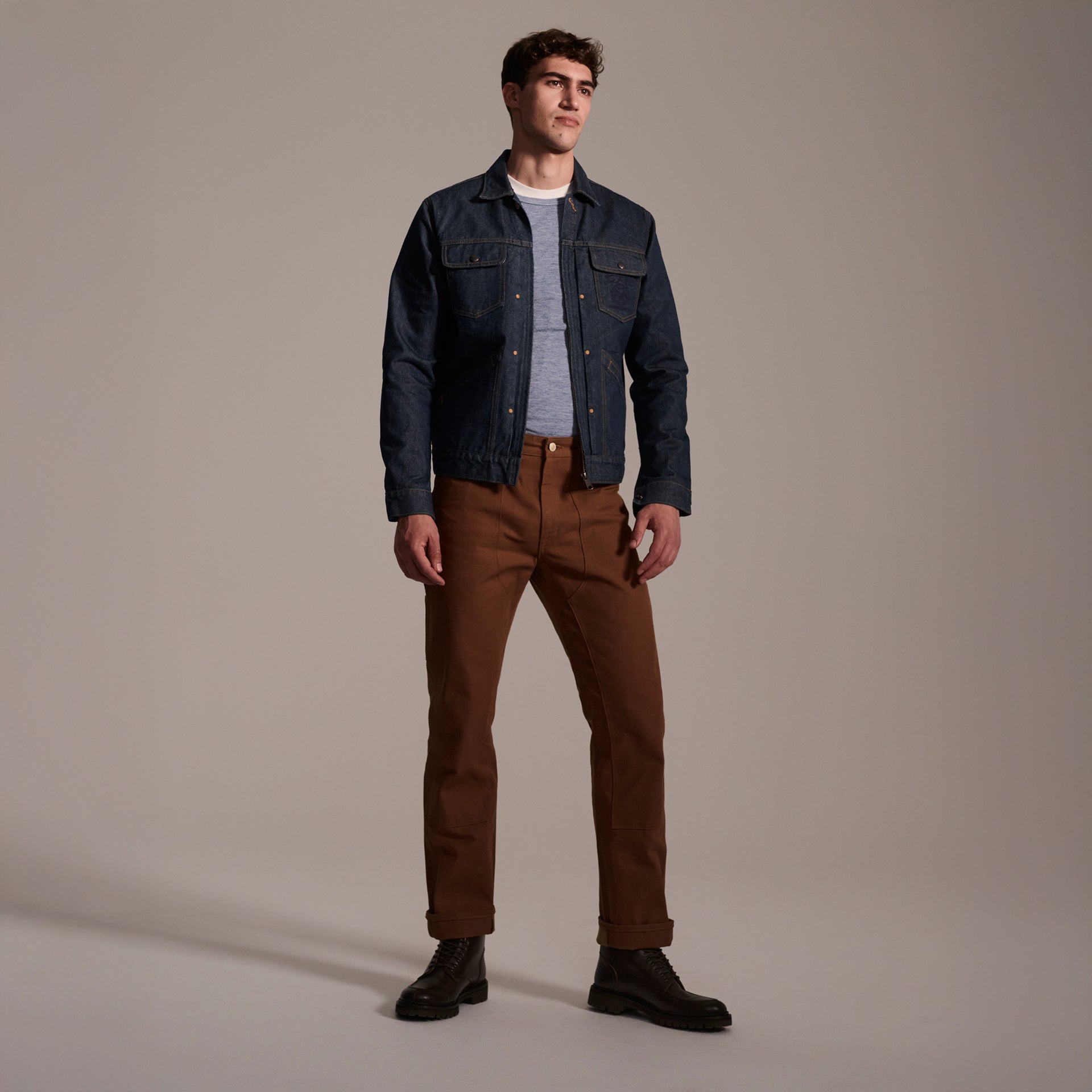 A man stands in a studio wearing a blue denim jacket, gray shirt, T7C The Seven Continents Sid Pant in Hide Raw, and black boots against a plain neutral background.