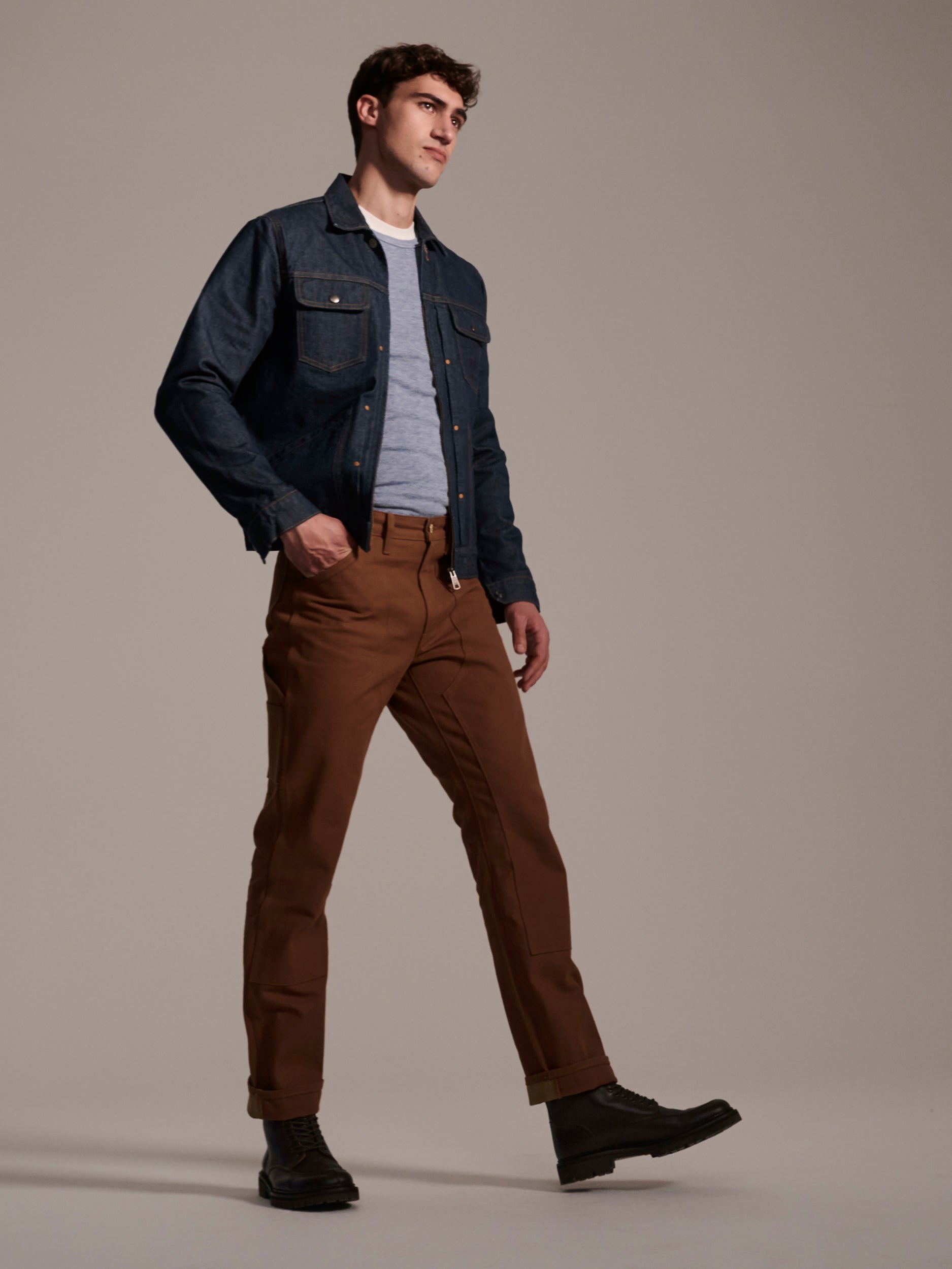 A man in a denim jacket, gray tee, and T7C The Seven Continents The Sid Pant in Hide Raw strides in black boots against a plain beige background.