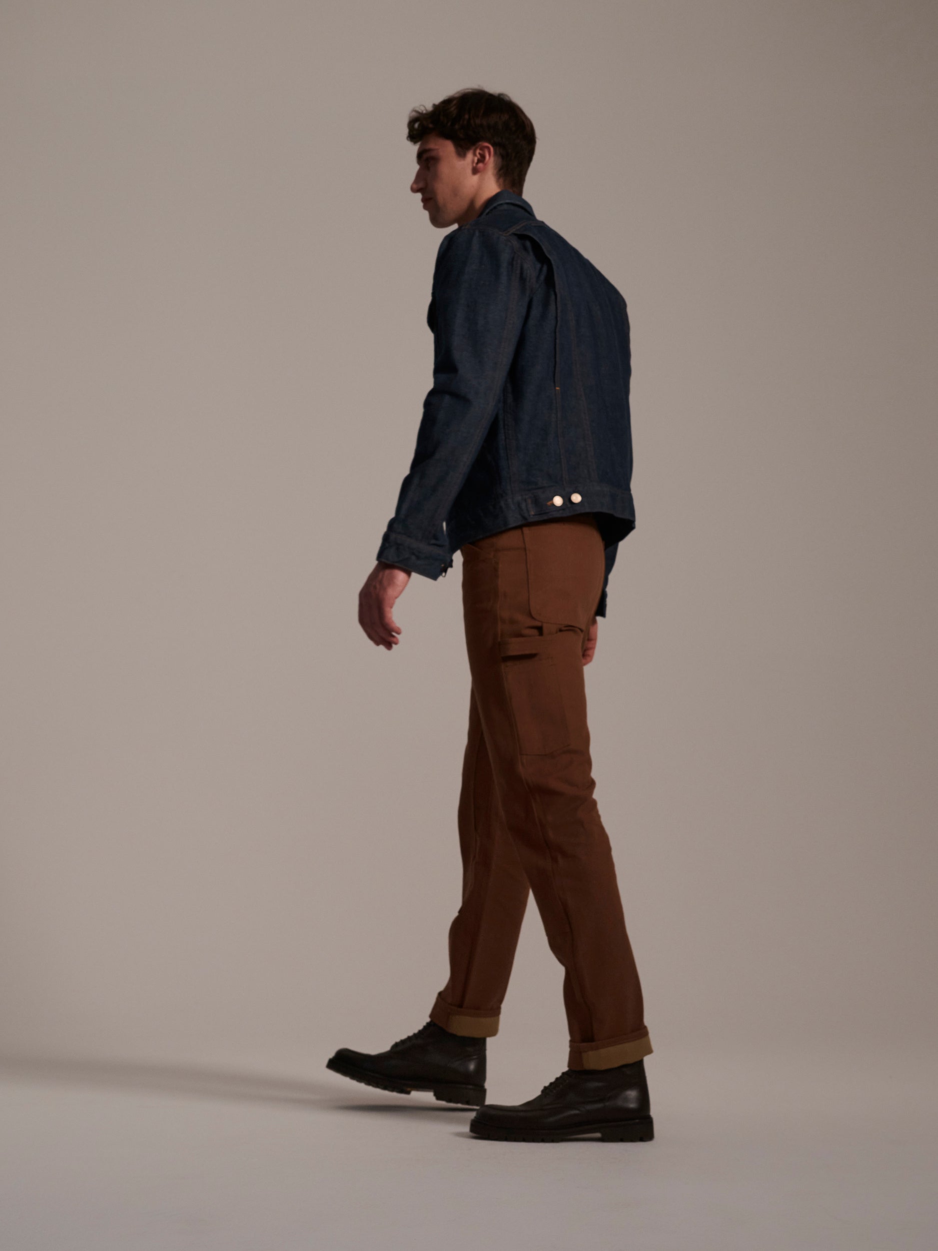 A man walks left against a light background, wearing a blue denim jacket, black shoes, and T7C The Seven Continents’ The Sid Pant in Hide Raw for a classic workwear look.
