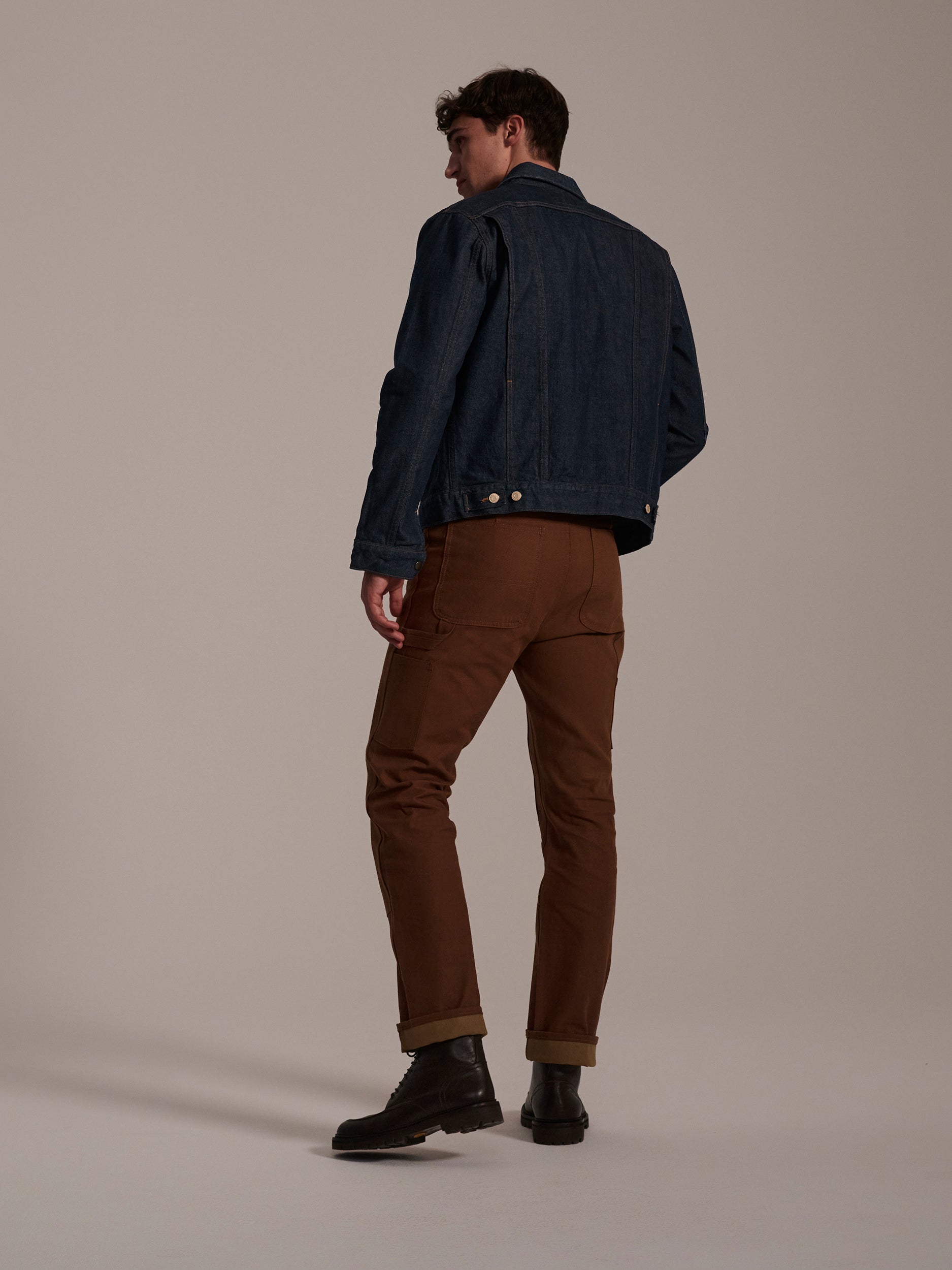 A man faces away, wearing a dark denim jacket, dark boots, and T7C The Seven Continents Sid Pant in Hide Raw. He stands against a plain, light-colored background.