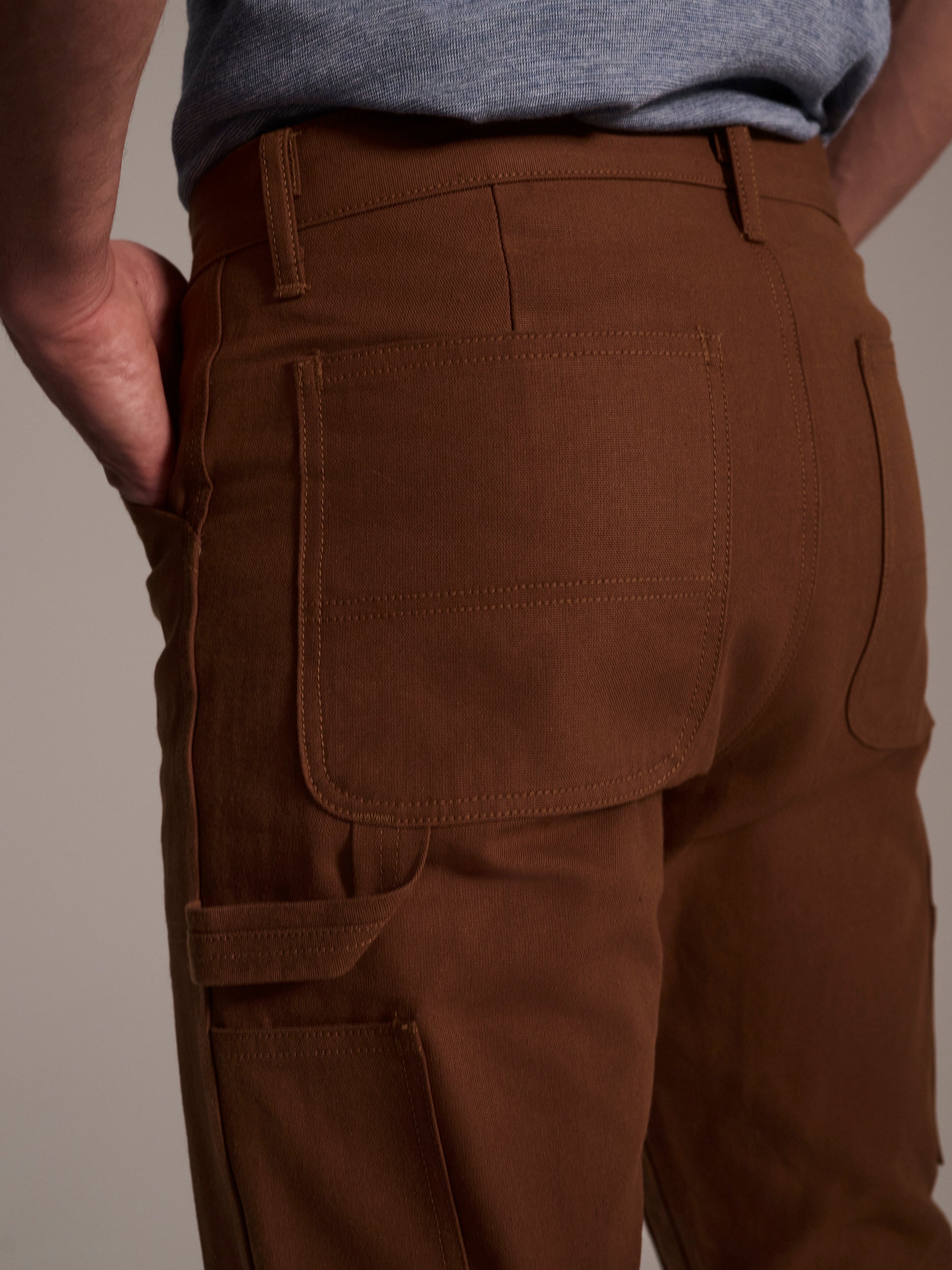 A close-up of someone wearing T7C The Seven Continents’ The Sid Pant in Hide Raw, featuring large back pockets and a hand in the side pocket, paired with a gray shirt—perfect for functional workwear.