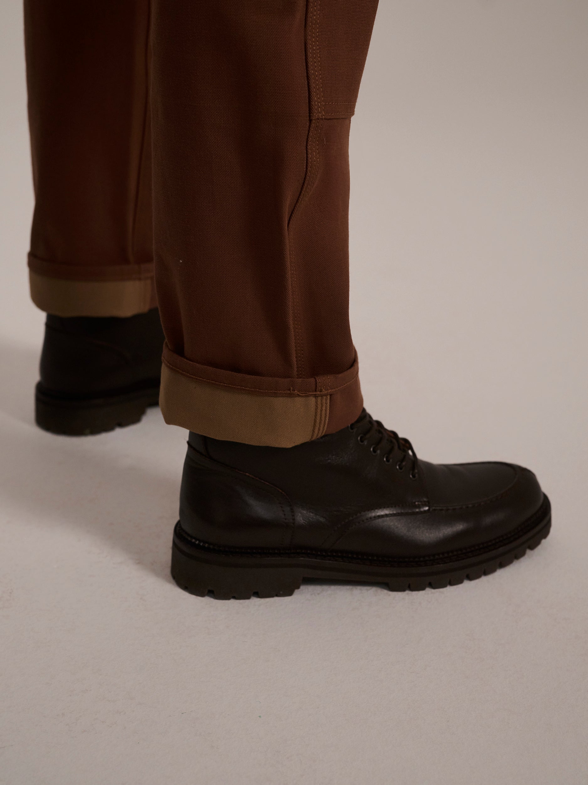 A person wearing T7C The Seven Continents The Sid Pant in Hide Raw and black leather lace-up boots stands on a light-colored surface.