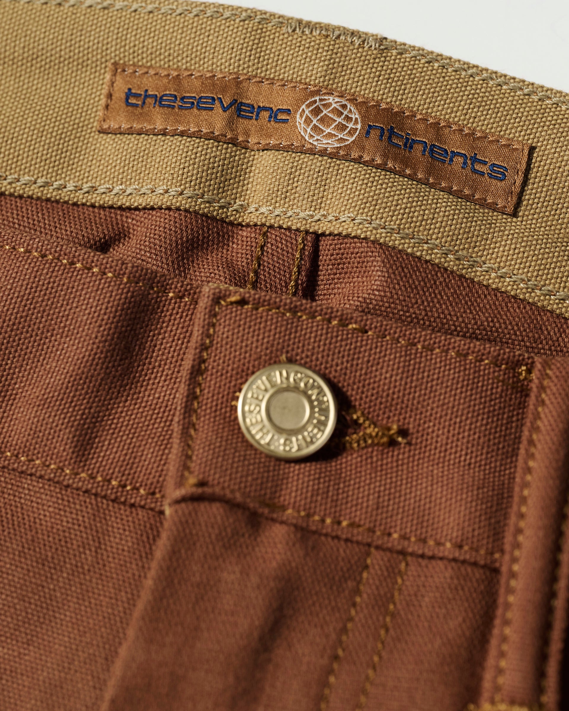 Close-up of The Sid Pant in Hide Raw by T7C The Seven Continents, featuring brown slim-straight legs, metal button, beige waistband, and a navy label with a globe icon—modern details on classic workwear.