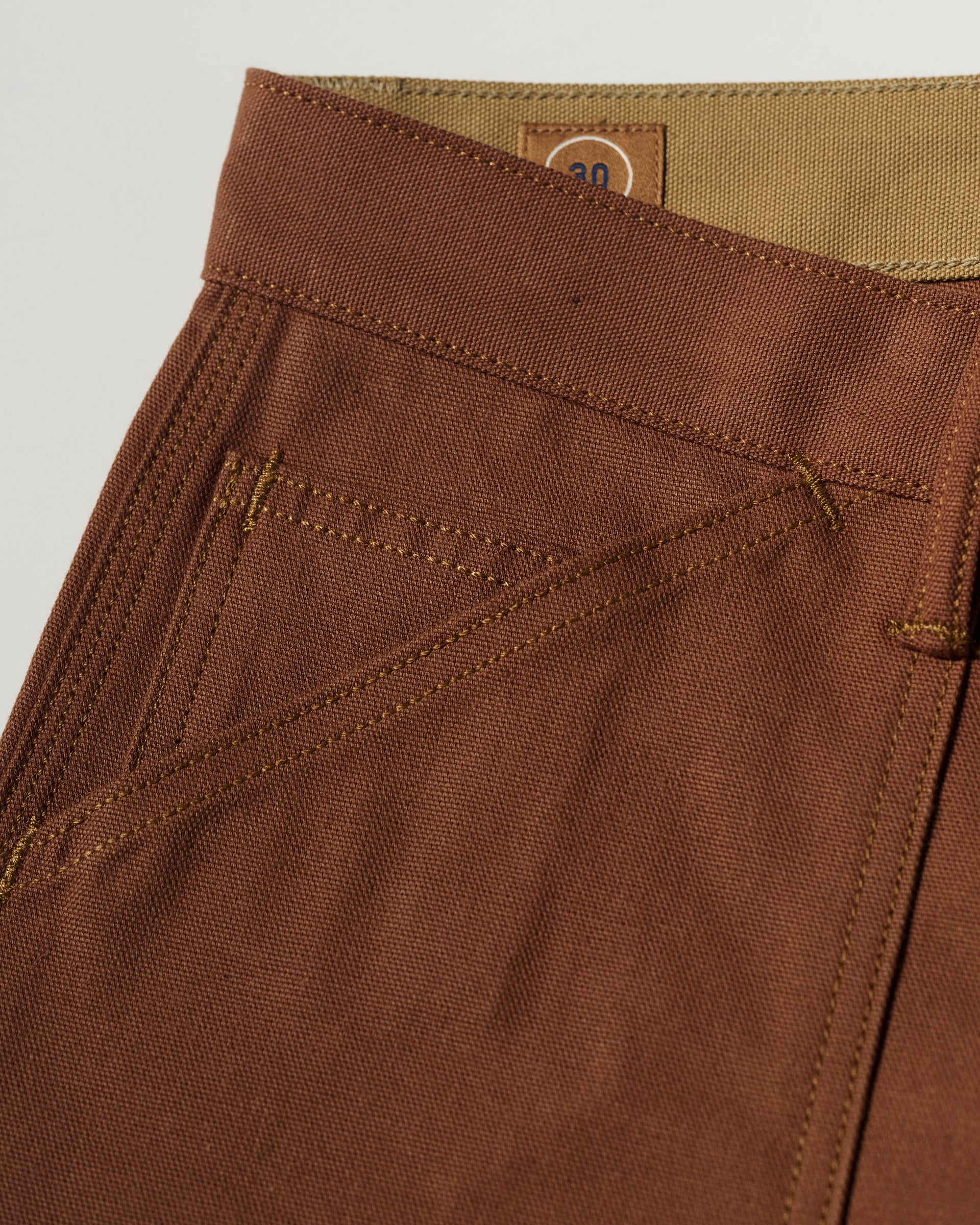 Close-up of T7C The Seven Continents The Sid Pant in Hide Raw—slim-straight brown pants featuring detailed stitching, front pocket, belt loop, and a small tan 30 label at the waistband for classic workwear style with refined utility.