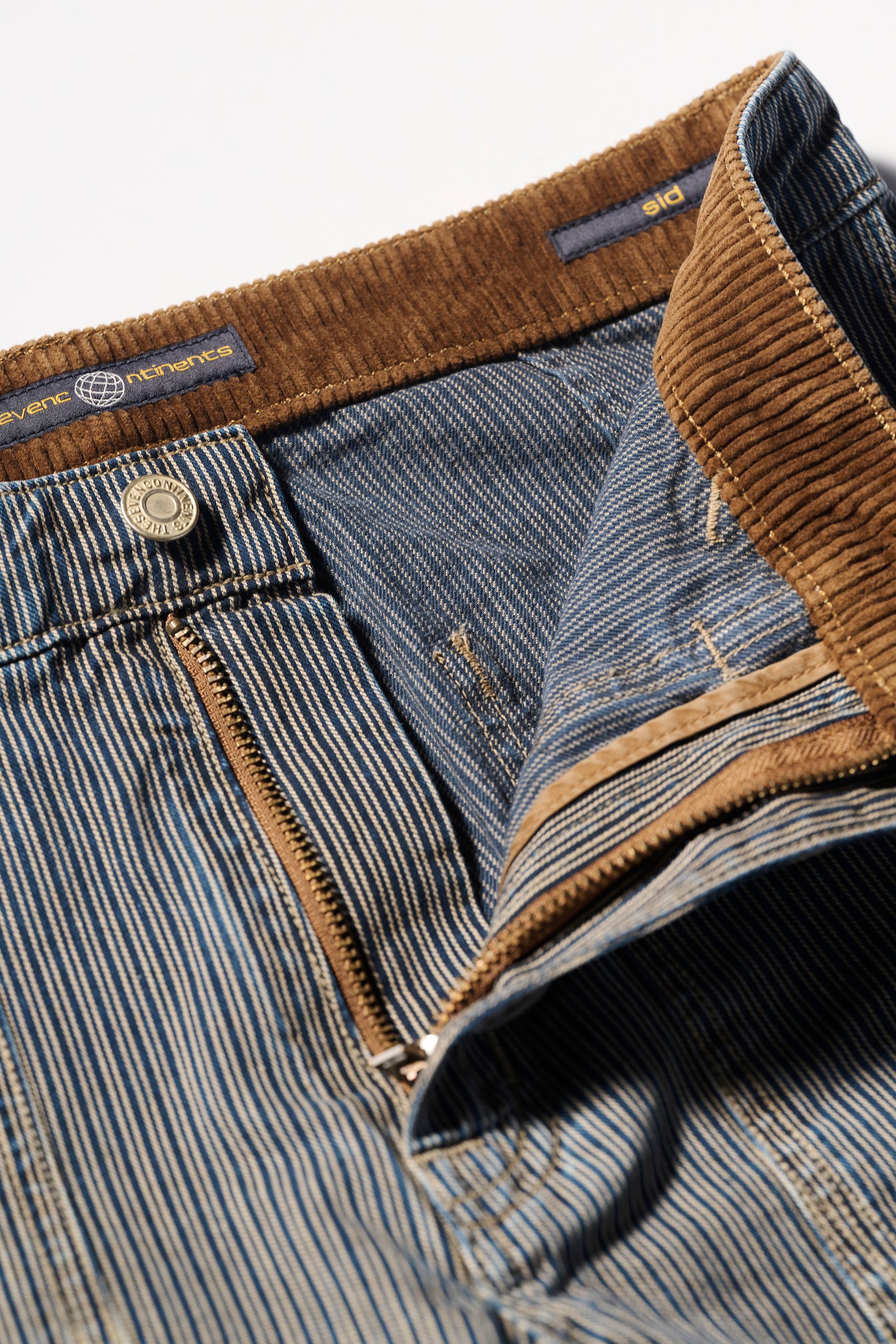 Close-up of The Sid Pant in Indigo Stripe by T7C The Seven Continents, featuring indigo railroad stripe cotton, a brown corduroy waistband, partly open zipper, and a metal waist button.