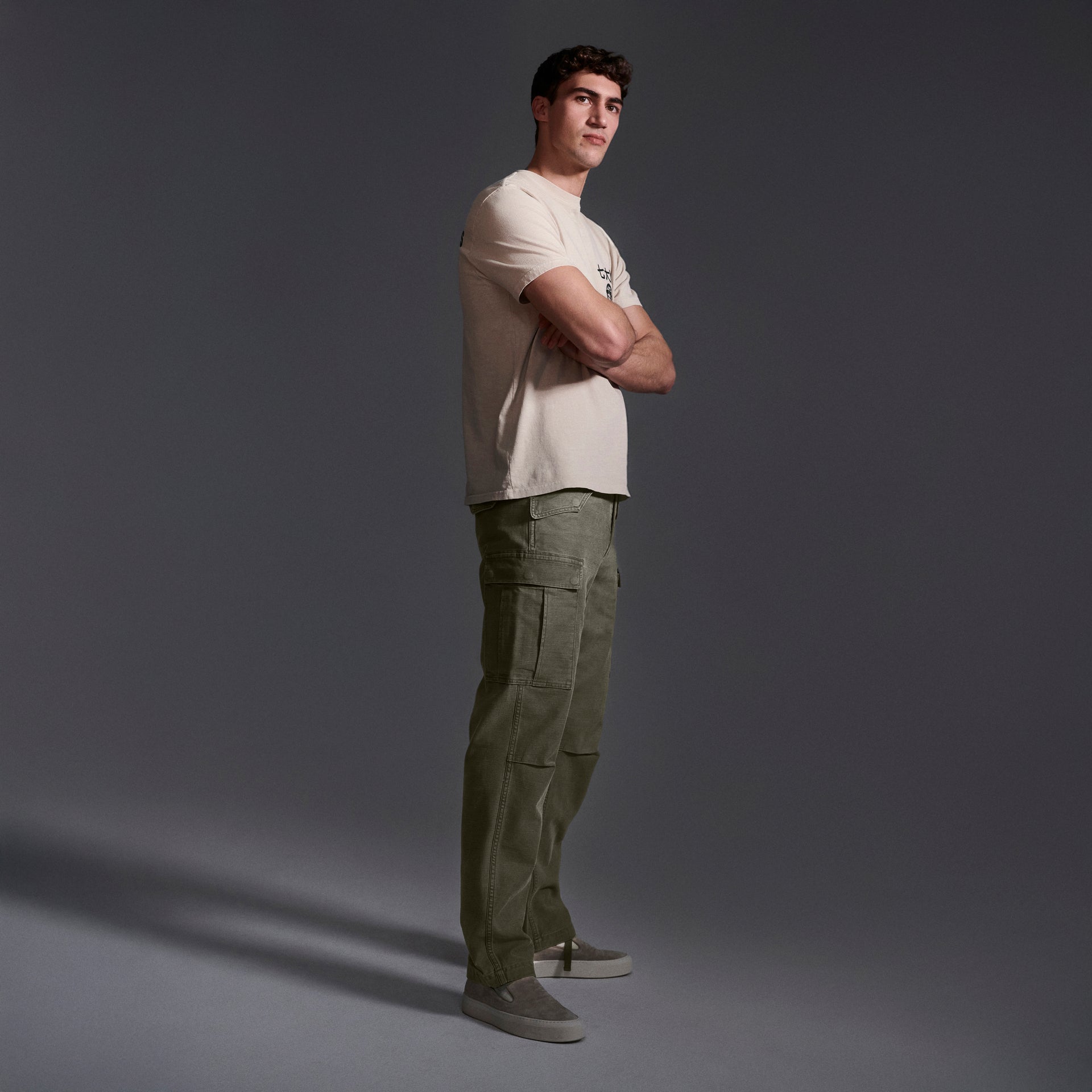 A man stands sideways with arms crossed, wearing a beige t-shirt and T7Cs The Brando Cargo in Olive Drab, plus gray slip-on shoes, against a dark gradient background.