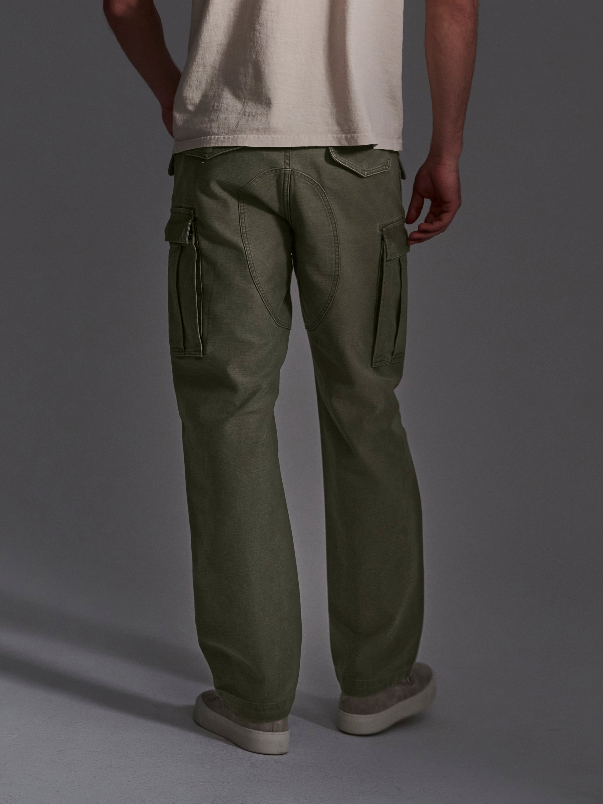 A person stands with their back to the camera against a gray background, wearing T7Cs The Brando Cargo in Olive Drab, a loose-fitting cotton-back satin pant, paired with a cream T-shirt and white sneakers.
