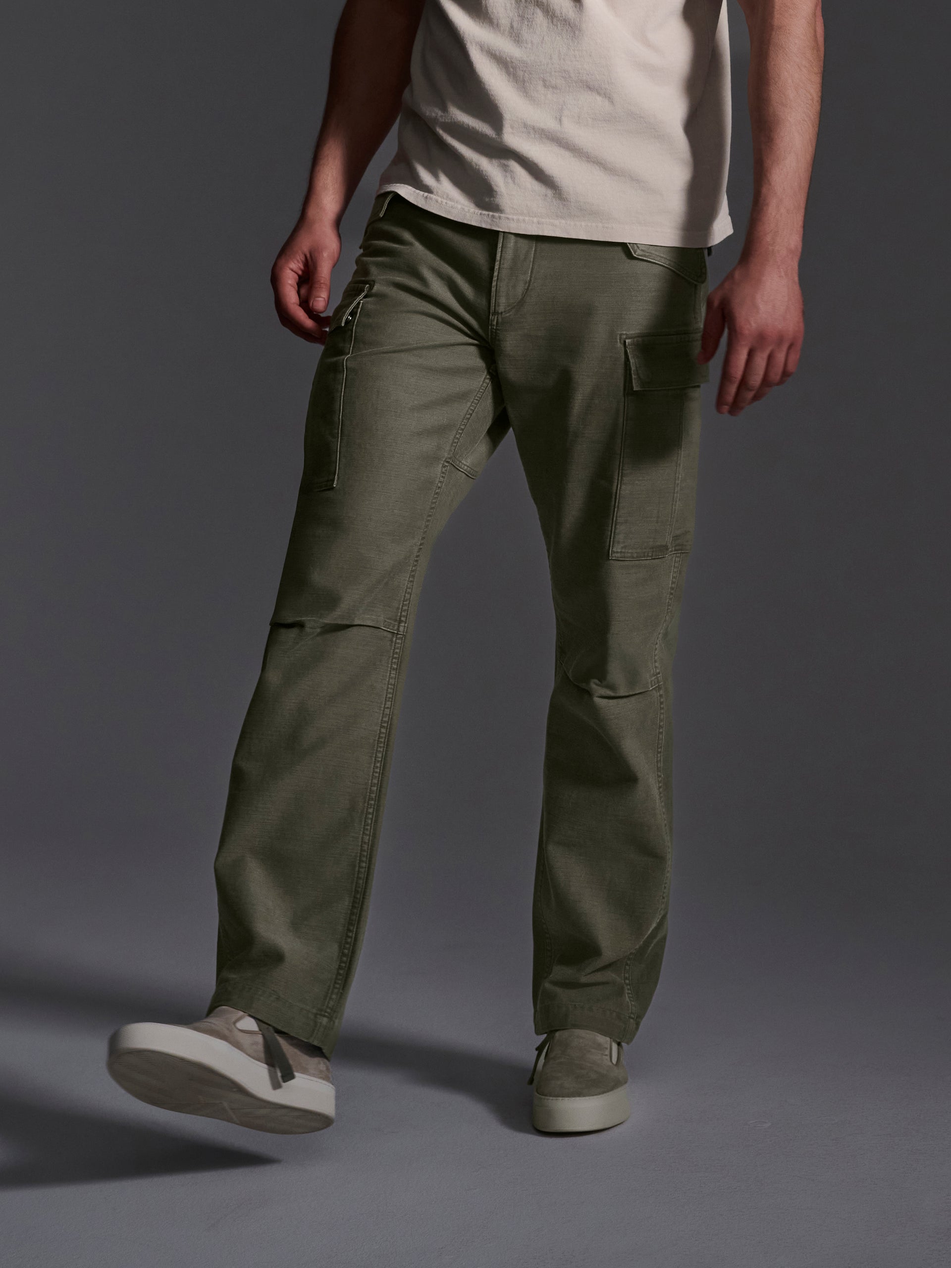 A person models the T7C Brando Cargo in Olive Drab with a light beige tee and gray sneakers, standing against a dark background, their face out of frame.