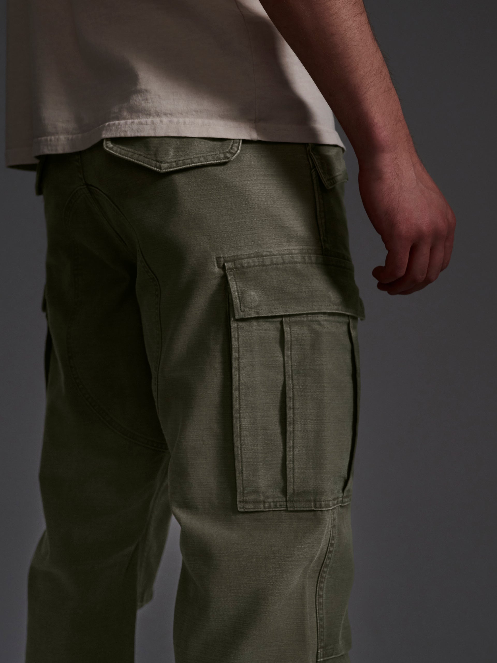 A person stands sideways against a dark background, wearing a light beige t-shirt paired with T7Cs The Brando Cargo in Olive Drab, with their right arm relaxed by their side.