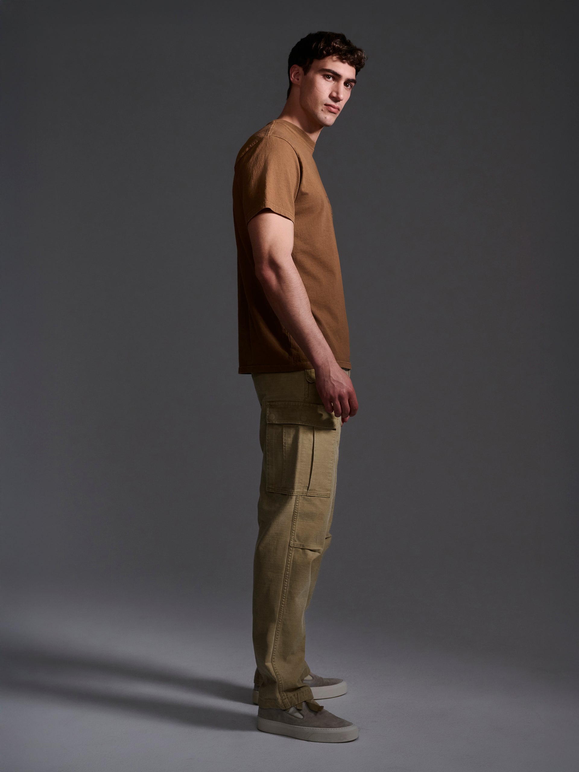 A man in profile against a dark background wears The Brando Cargo in Sand by T7C, a brown cotton-back satin T-shirt, and gray sneakers. He looks slightly toward the camera with a neutral expression.