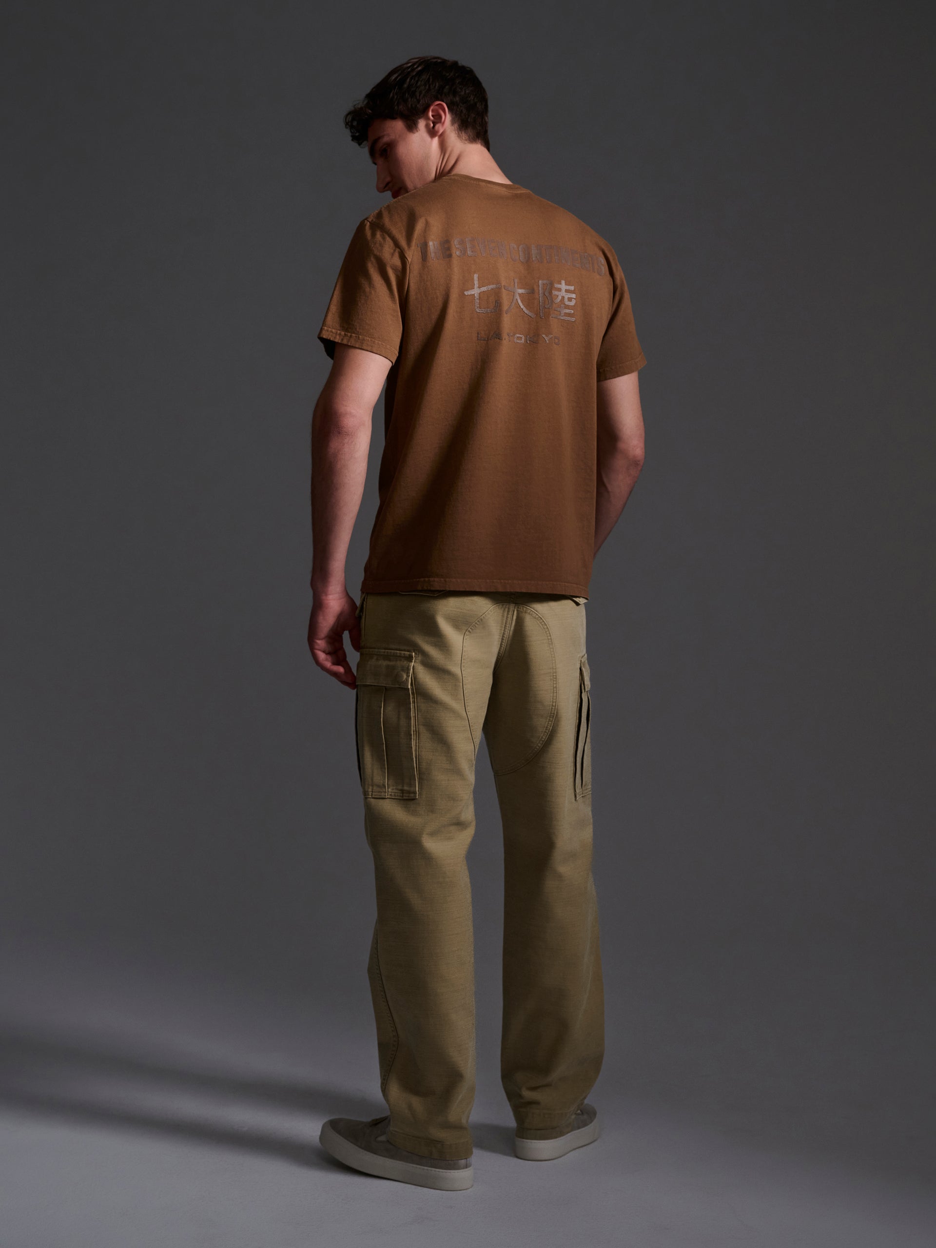 A man stands facing away, wearing a brown sulphur-dyed T-shirt with white back text and T7Cs The Brando Cargo in Sand pants. The background is dark, and his head is turned slightly left.