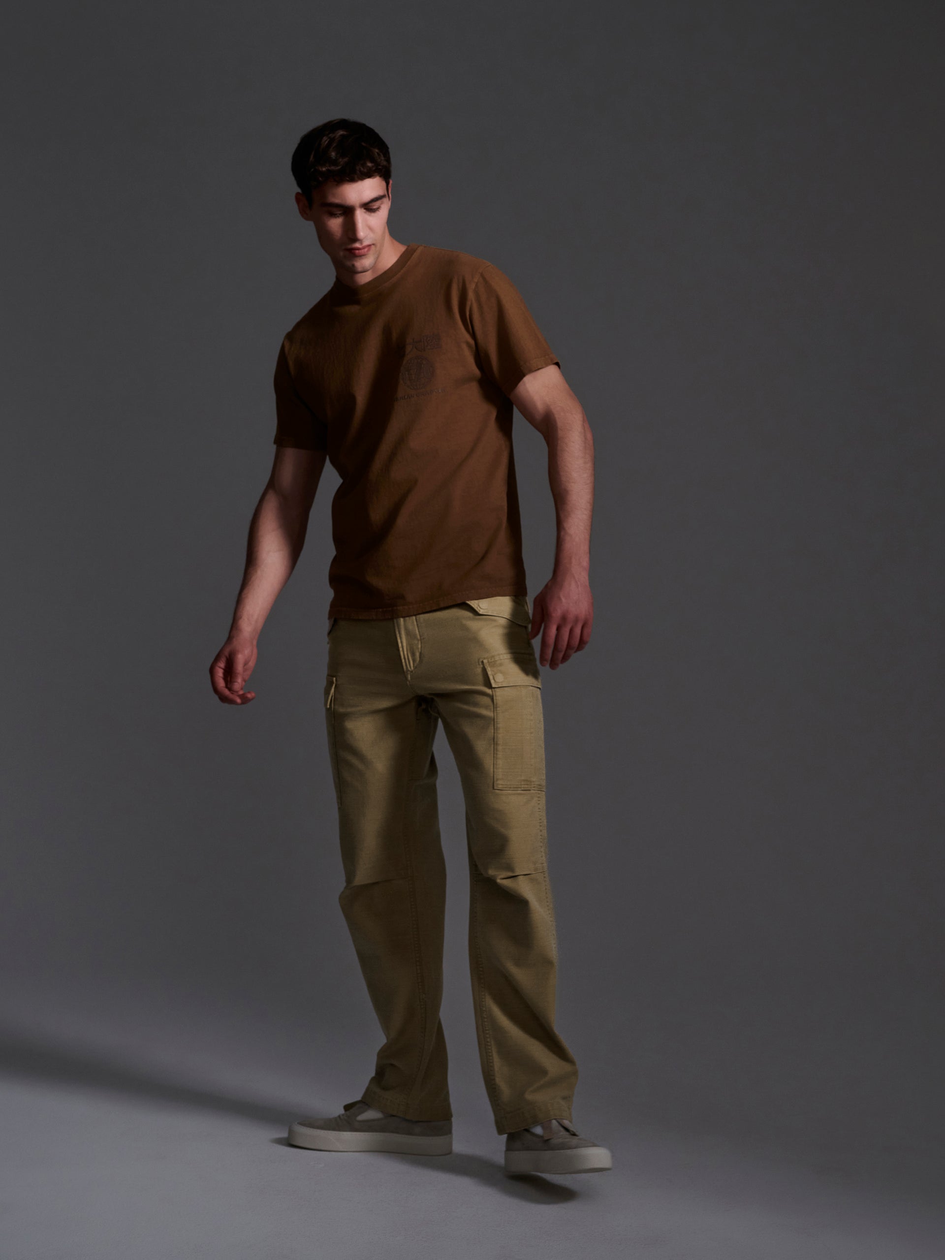 A man stands against a gray background wearing The Brando Cargo in Sand by T7C, crafted from cotton-back satin. He looks down to his left, one hand by his side and the other slightly bent.