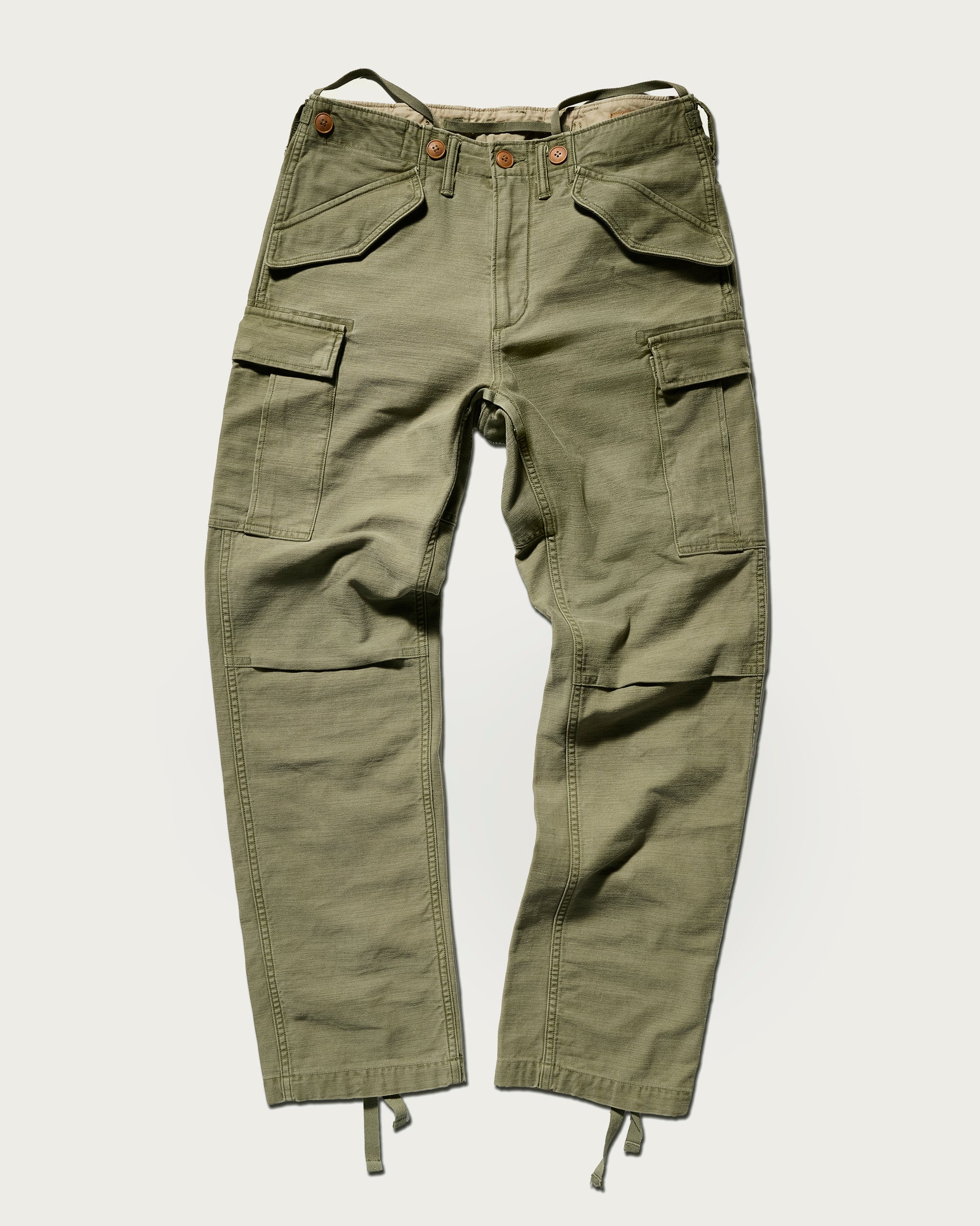 The Brando Cargo in Olive Drab by T7C, crafted from cotton-back satin with a sulphur-dyed finish, features multiple pockets, button and zip closure, adjustable waist tabs, and cuff drawstrings—shown laid flat on a light background.