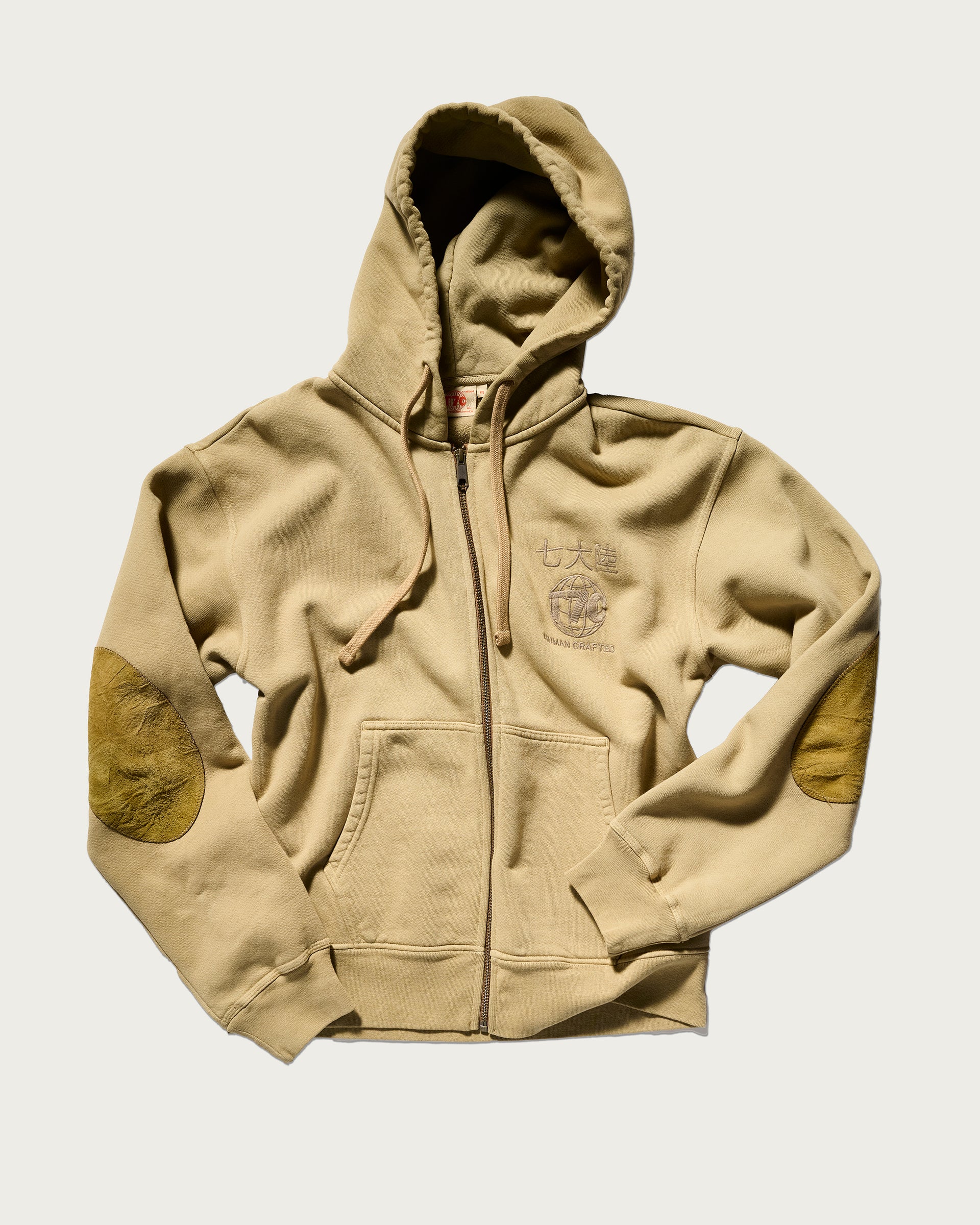 The Camden Hoodie in Desert by T7C The Seven Continents features a beige color, hood, drawstrings, front pockets, genuine suede elbow patches, and subtle left chest graphics, all shown flat on a light background.