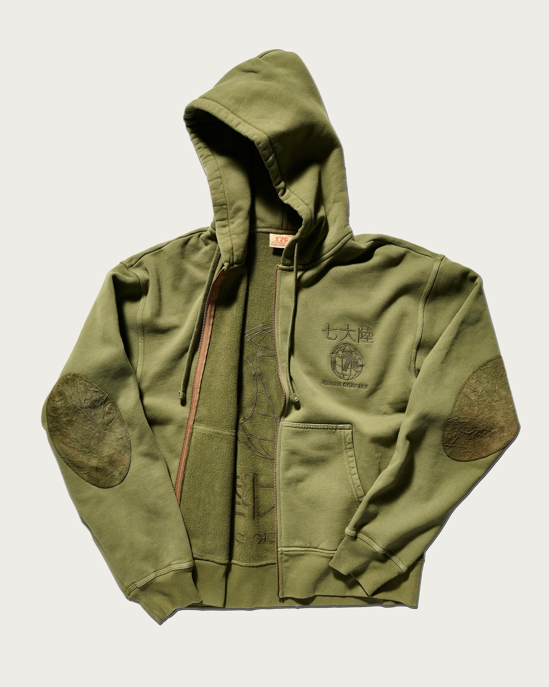The Camden Hoodie in Olive by T7C The Seven Continents features suede elbow patches, a front zipper, and Asian characters above a shield emblem on the left chest. The hoodie is displayed flat with one sleeve partially folded.