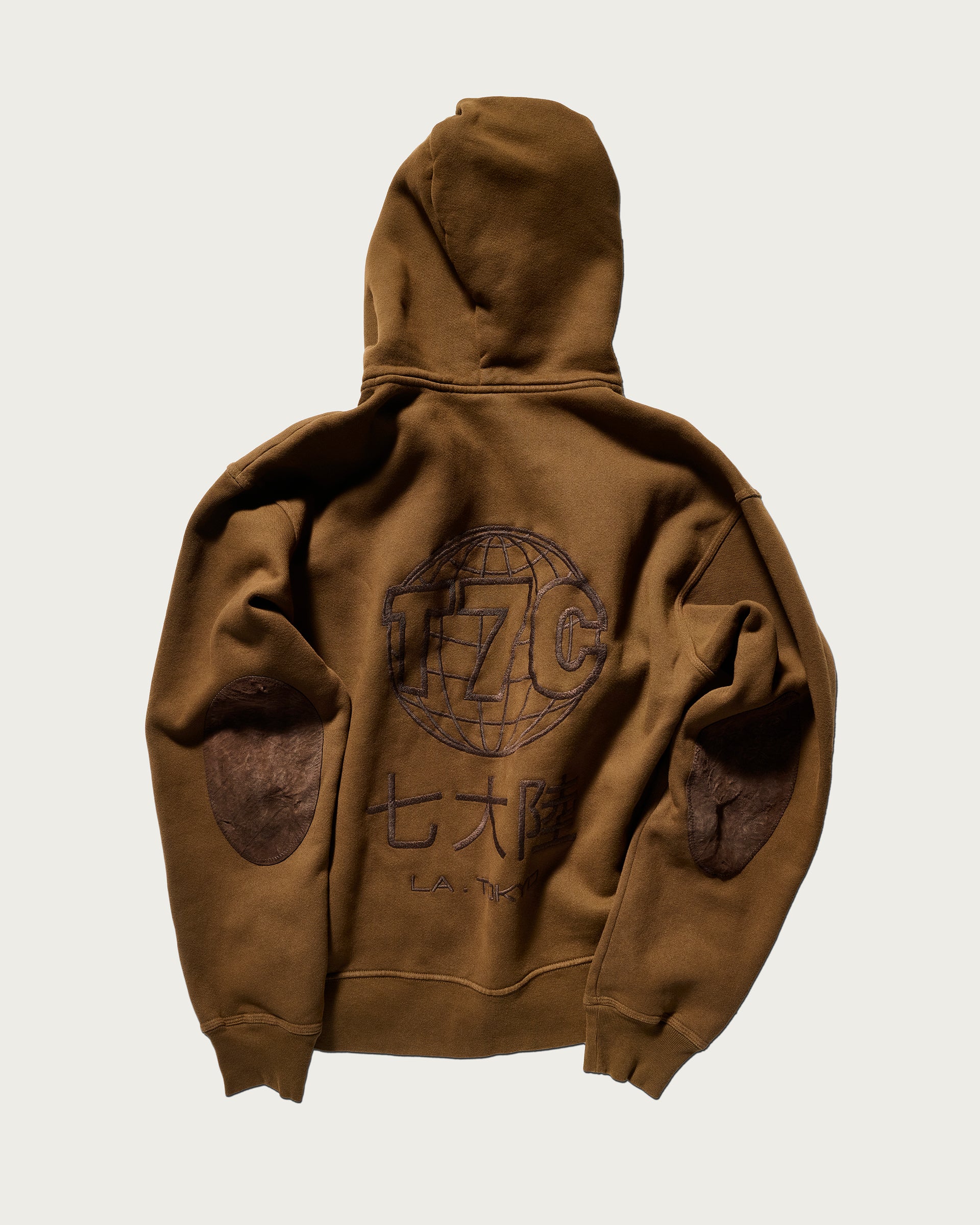 The Camden Hoodie in Pecan by T7C The Seven Continents features a relaxed fit, suede elbow patches, and an embroidered globe with “T7C,” Japanese characters, and “L.A. TOKYO” on the back. Shown laid flat on a light background.