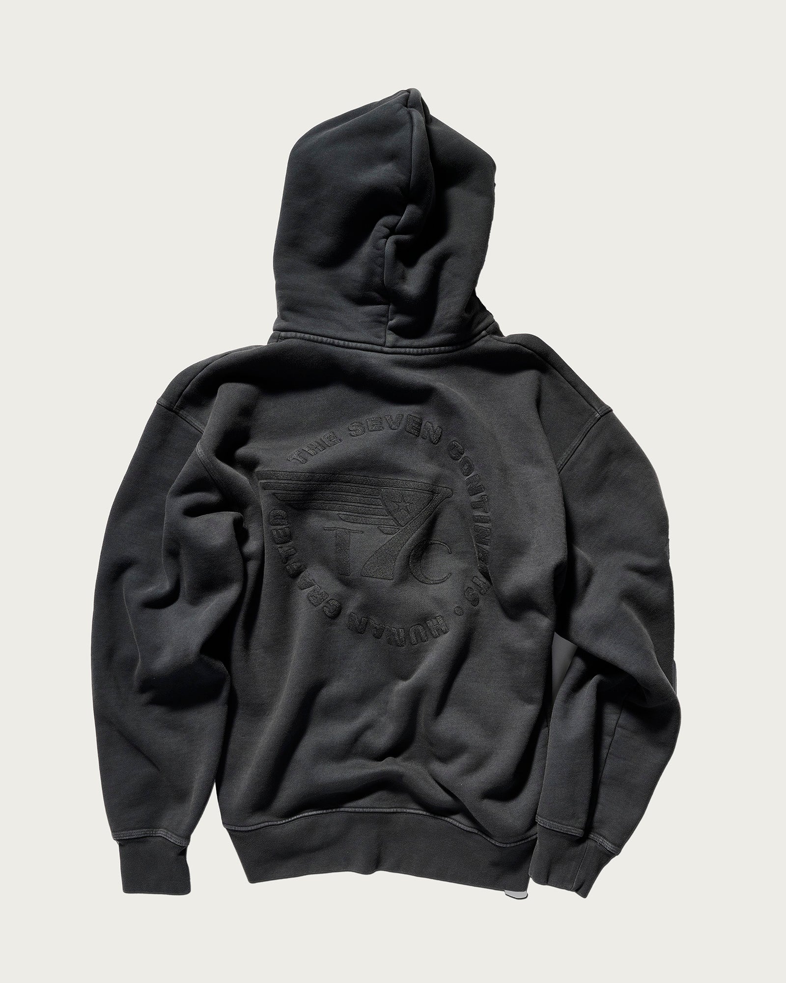 THE DAX IN VINTAGE BLACK by T7C is a loose-fit black hoodie, laid flat to show its back with an embossed circular logo, text, and a central outstretched hand design. Visible seams add detail to the overall look.