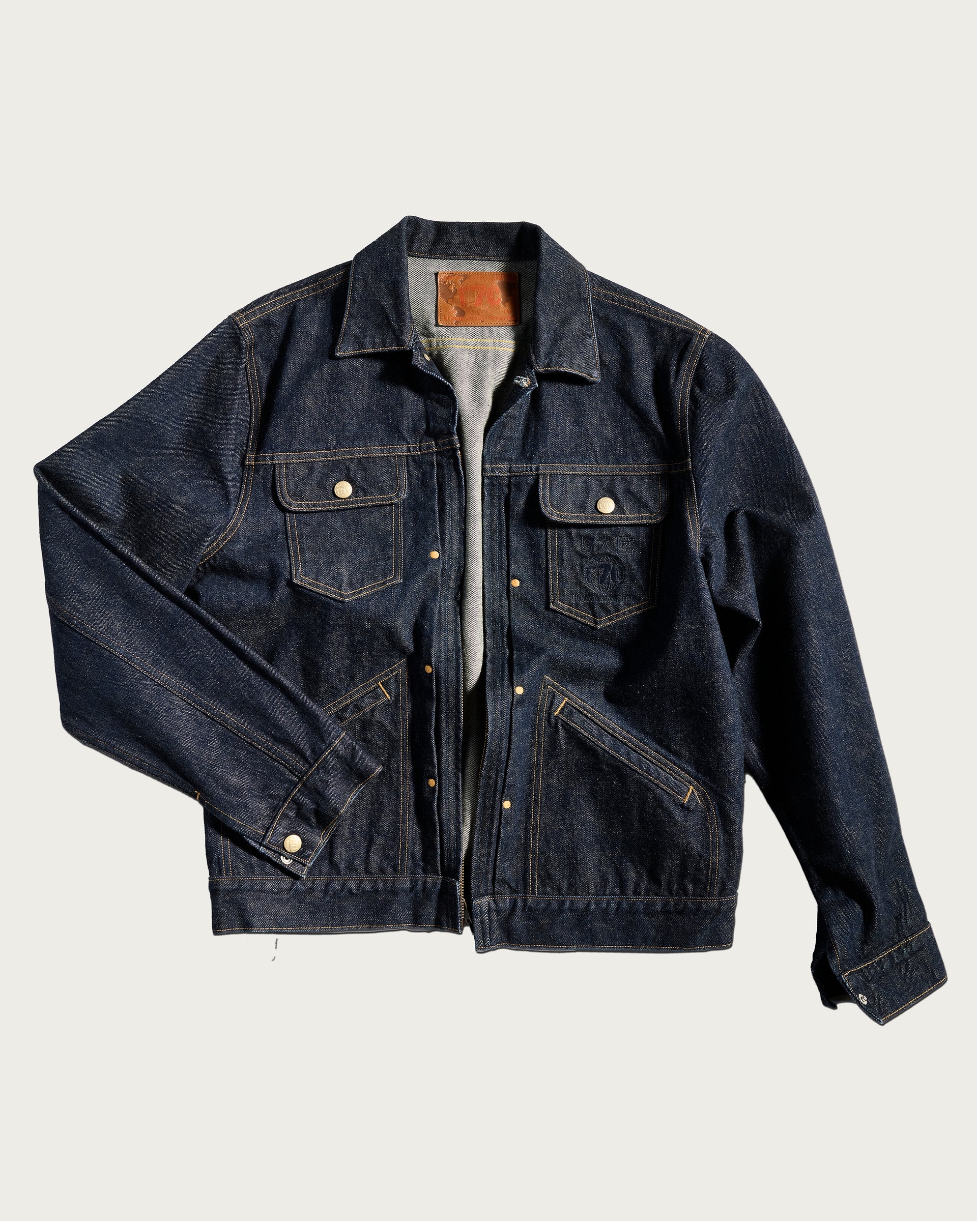 The T7C The Seven Continents Holden Classic Jacket with Zipper in dark blue denim features buttoned chest pockets, side pockets, tan stitching, and a subtle Japan Logo, laid flat on a light background with one sleeve slightly bent.