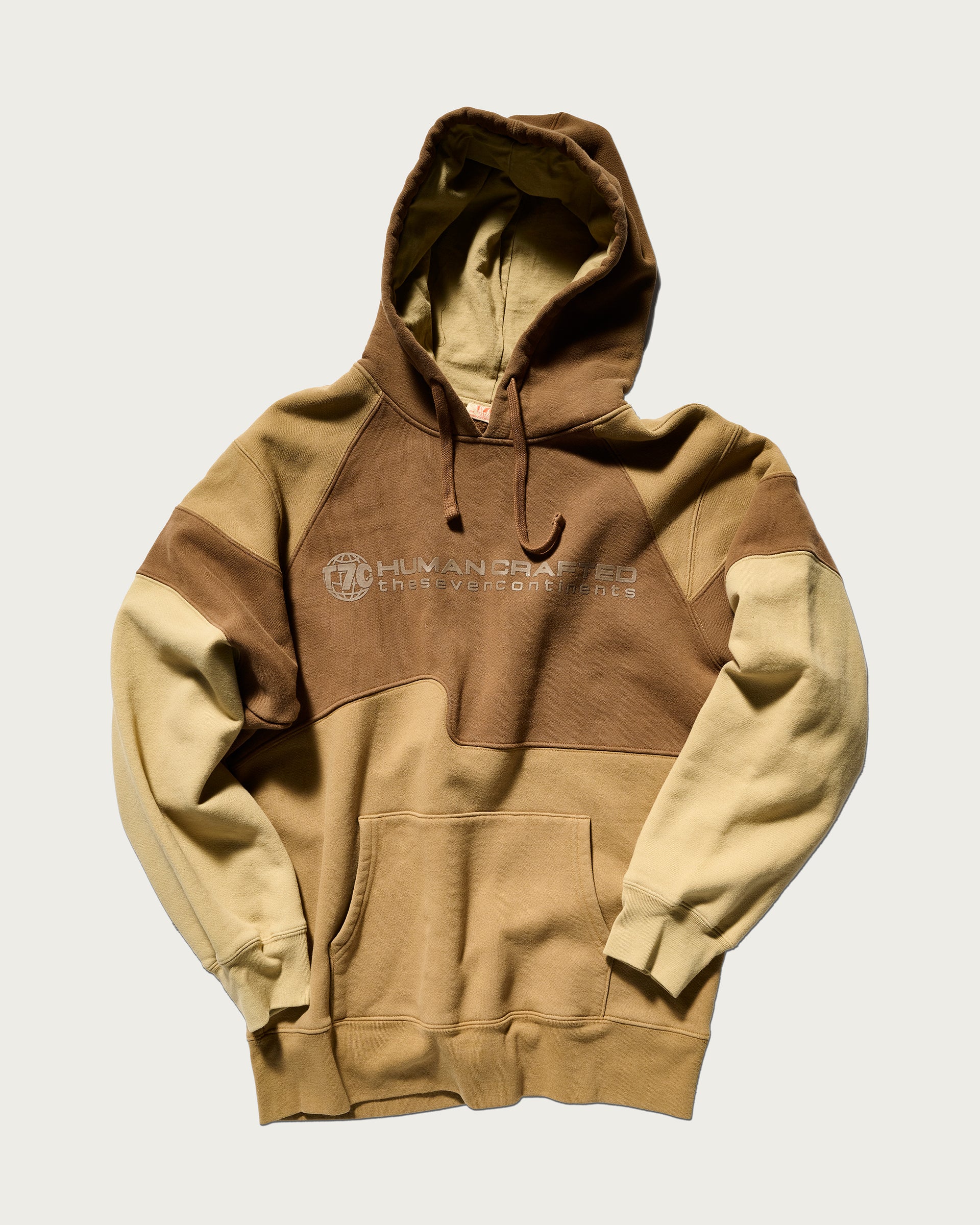 The Hunter Hoodie in Terra by T7C The Seven Continents features neutral color-blocking, a front pocket, hood, HUMAN DRAPED text on the chest, camo-inspired seams, and a subtle circular emblem.