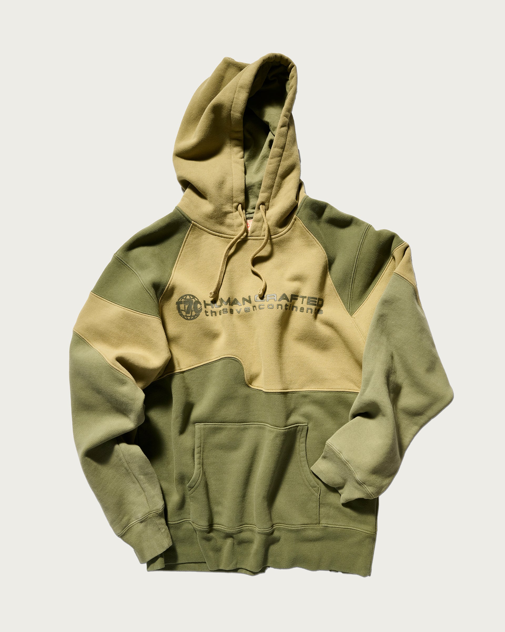 The Hunter Hoodie in Verde by T7C The Seven Continents features camo-inspired seams, a front pocket, drawstring hood, and HANDCRAFTED text with a globe design on the chest.