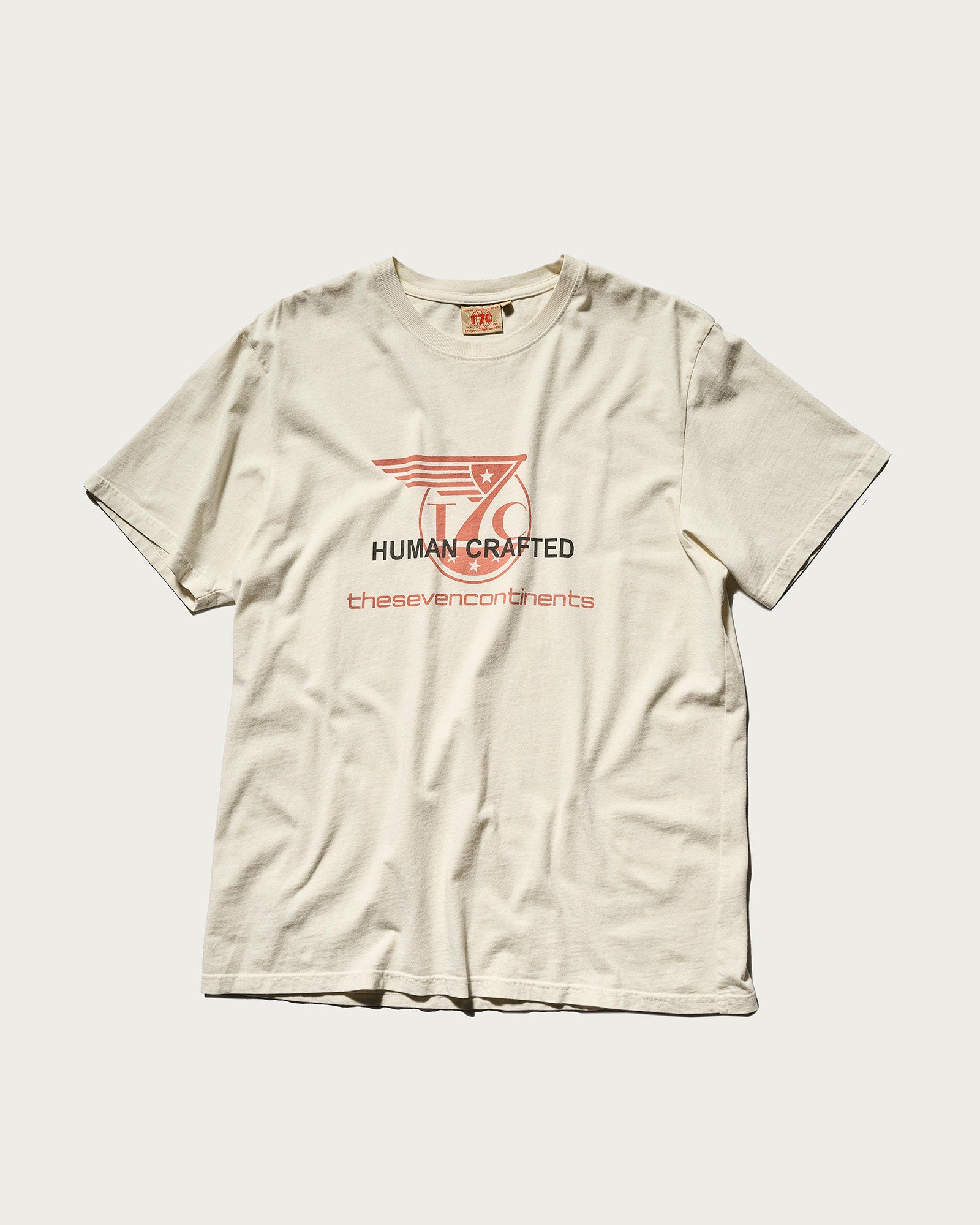 The cream-colored THE KNOXS TEE WITH ICON PRINT by T7C lies flat, featuring a red wing graphic and the text HUMAN CRAFTED and thesevencontinents on the front.