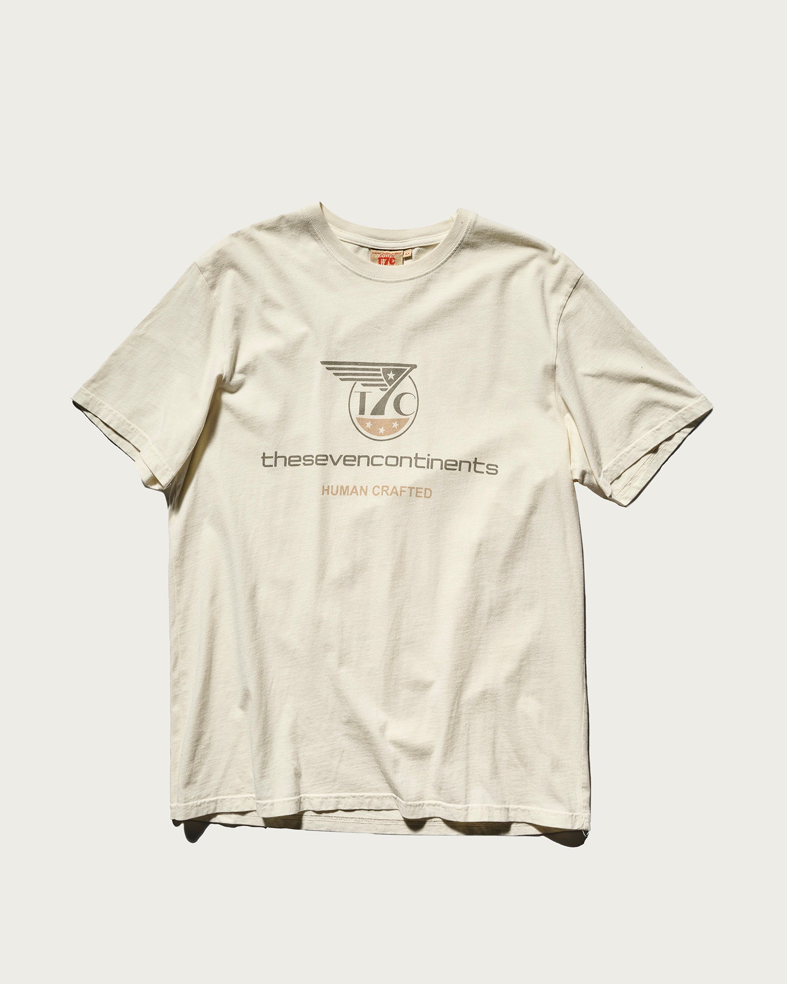 PRE-ORDER THE KNOXS TEE WITH STATION PRINT by T7C: a cream, stone washed tee made in Los Angeles, featuring the seven continents logo, stylized thesevencontinents text, and HUMAN CRAFTED on the front in gray and orange.