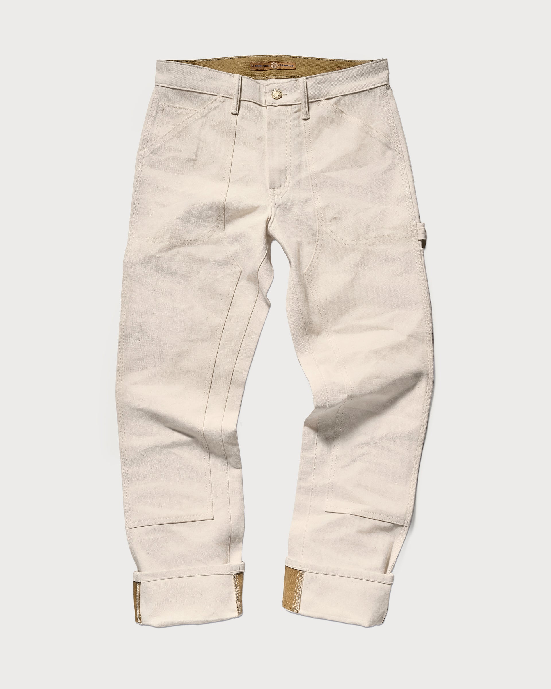 The Sid Pant in Chalk Raw by T7C The Seven Continents features reinforced stitching, rolled cuffs with brown lining, front and side pockets, and a hammer loop in a slim-straight workwear style, shown flat on a white background.