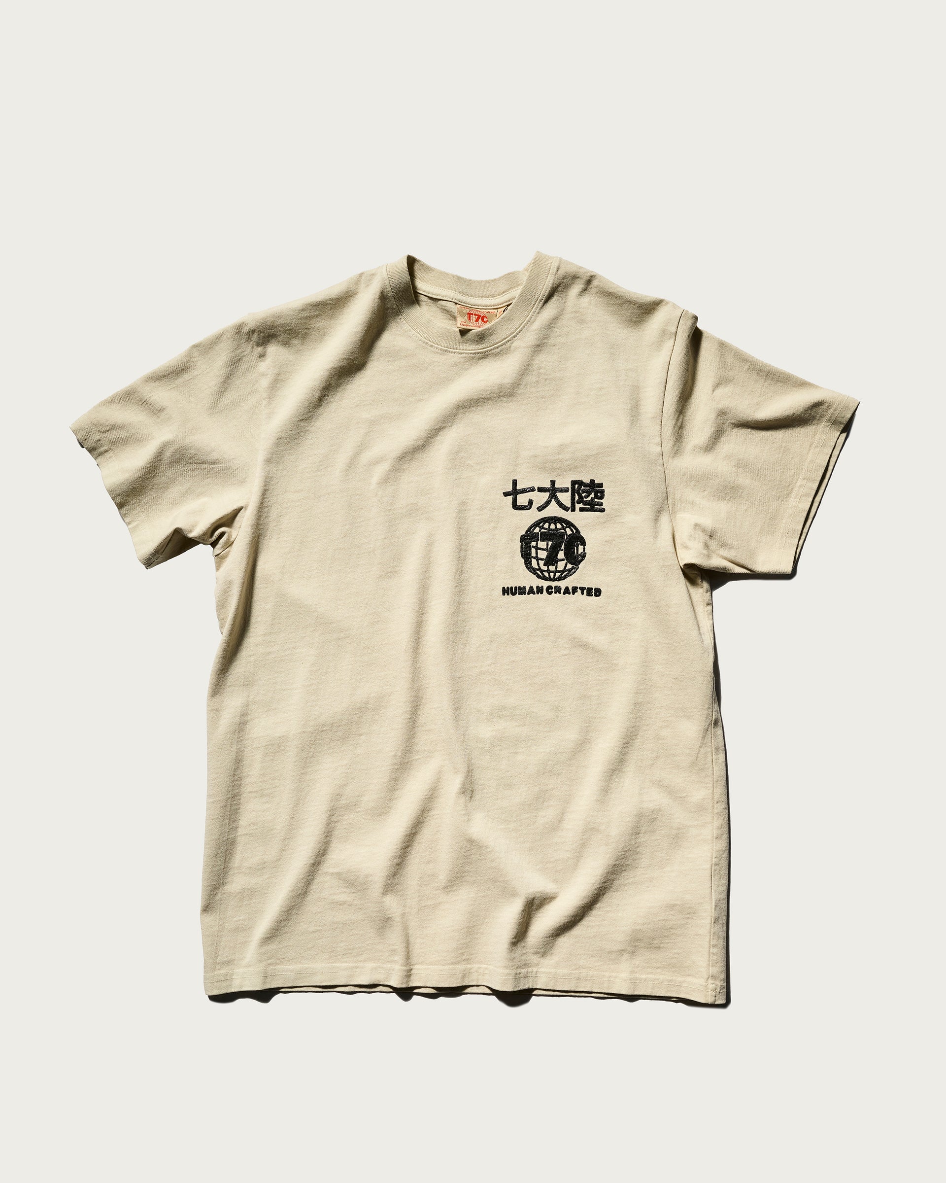 The Timo Tee in Fog by T7C The Seven Continents is a beige, textured shirt with black Japanese text, a globe graphic, and a puff screen printed HUMAN CRAFTED logo on the left chest, shown on a plain white background.