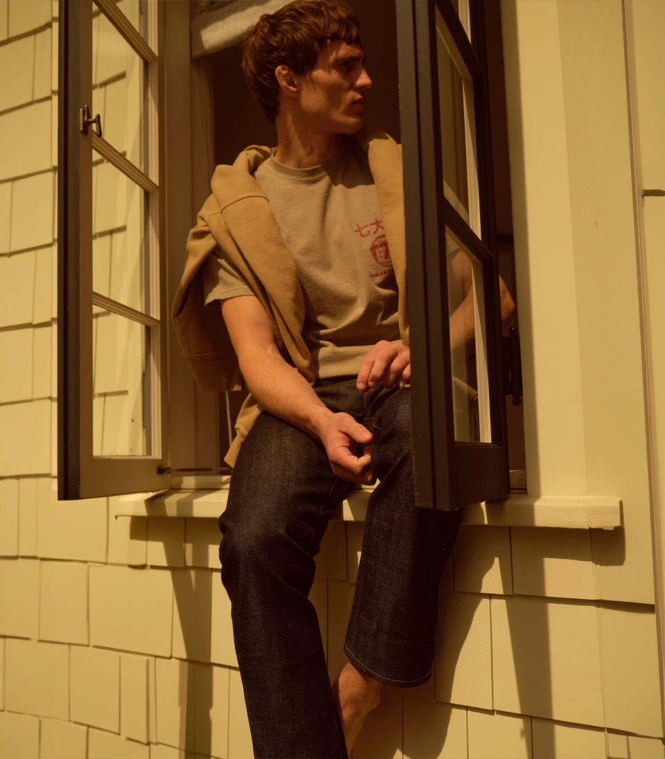 A male model wearing a t-shirt and jeans sitting on a window sill